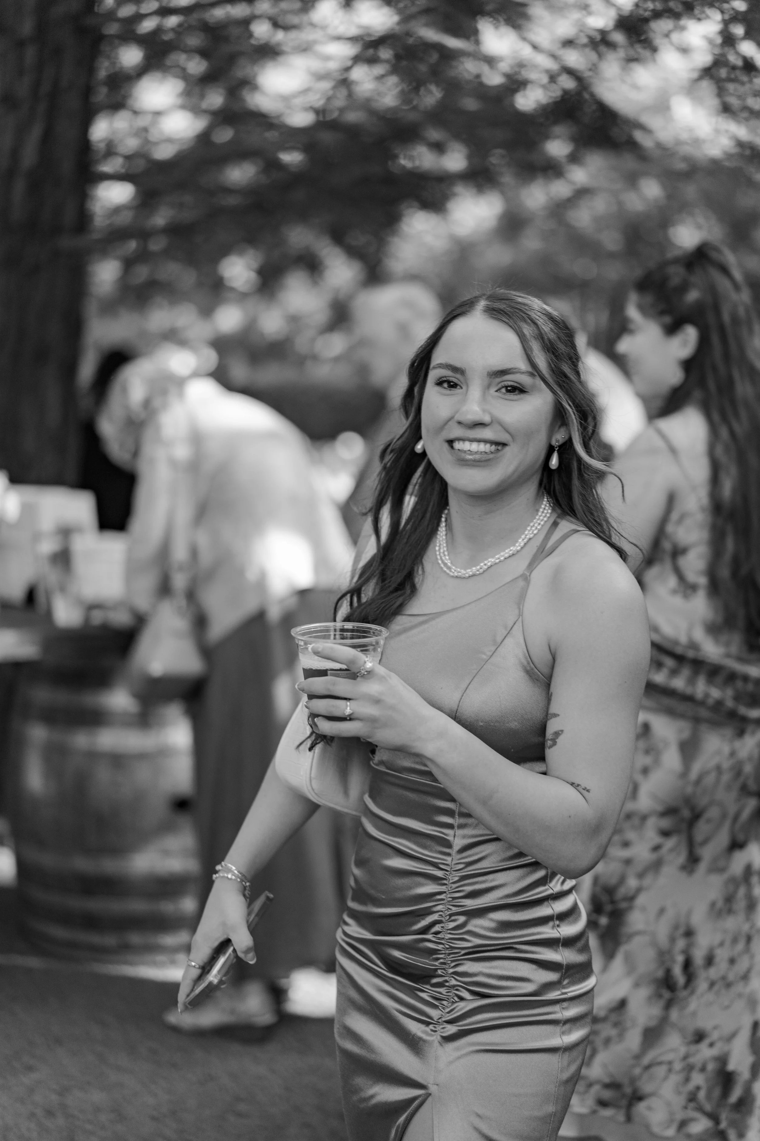 A woman smiling and holding a drink at an outdoor social event, wearing a satin dress and jewelry, with other people in the background.