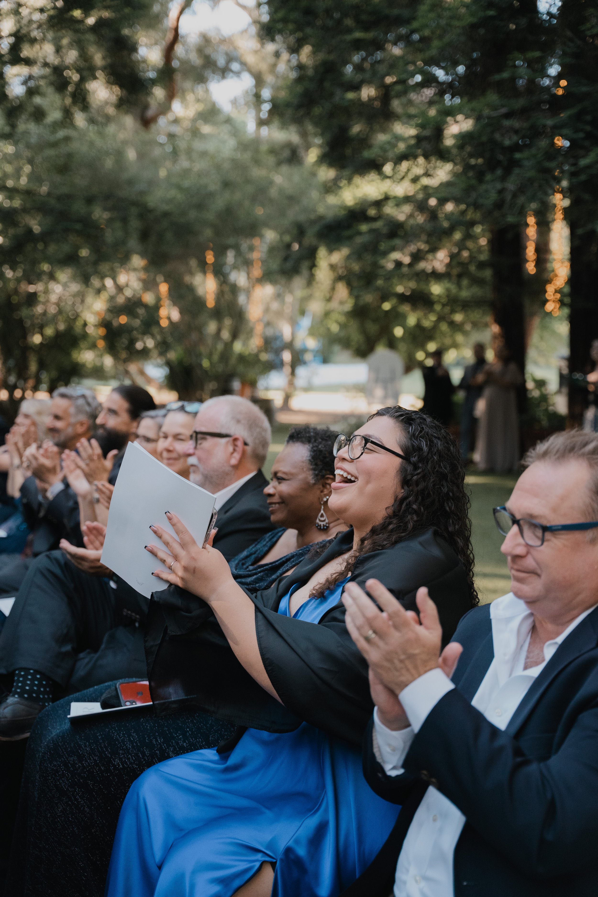 Group of people sitting outdoors, clapping and smiling, attending an event or celebration during the daytime, amidst trees and string lights.