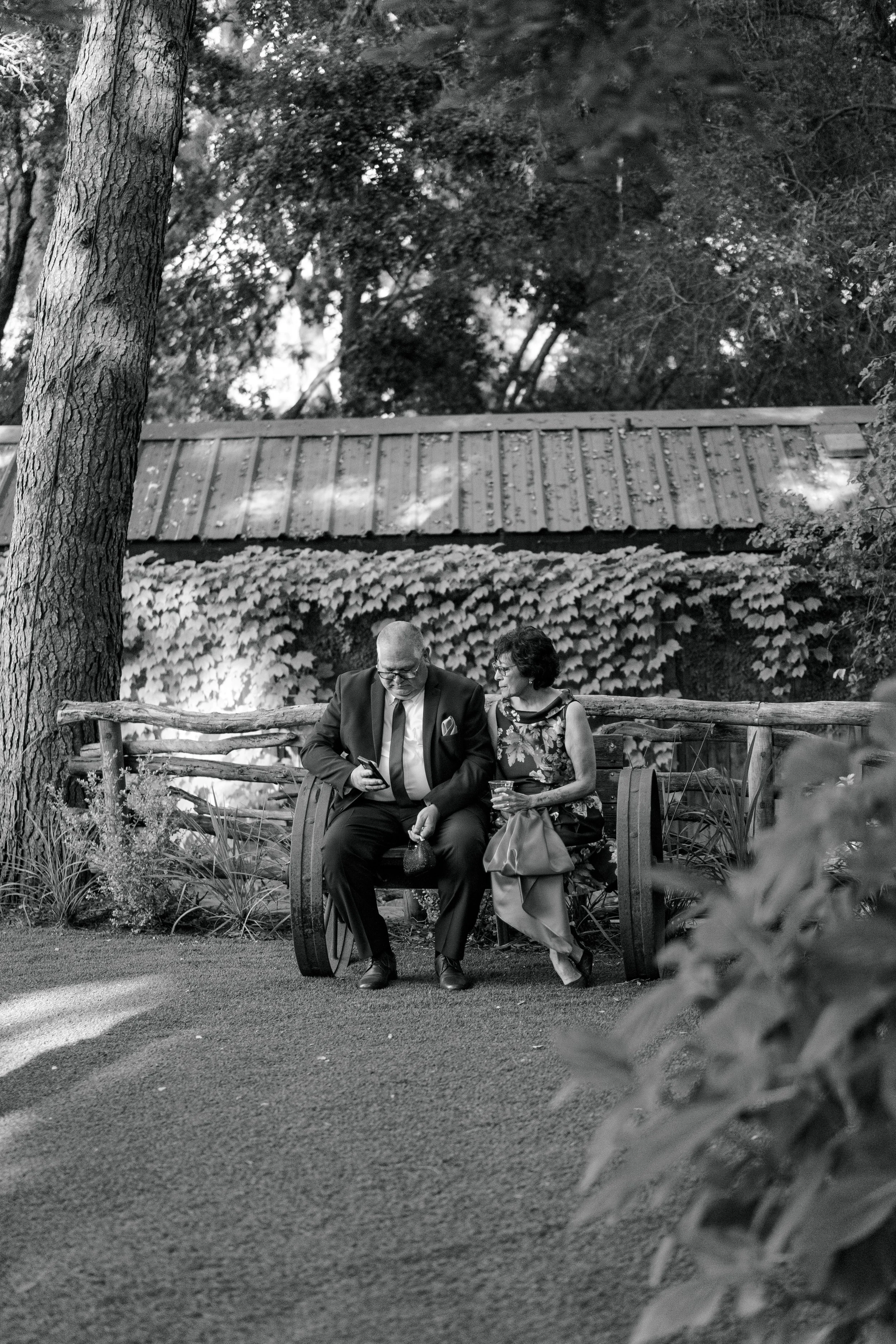 A man and a woman sitting on a bench in a park, engaged in a conversation. The man is wearing a suit and the woman a patterned dress, both appearing to be middle-aged or older.