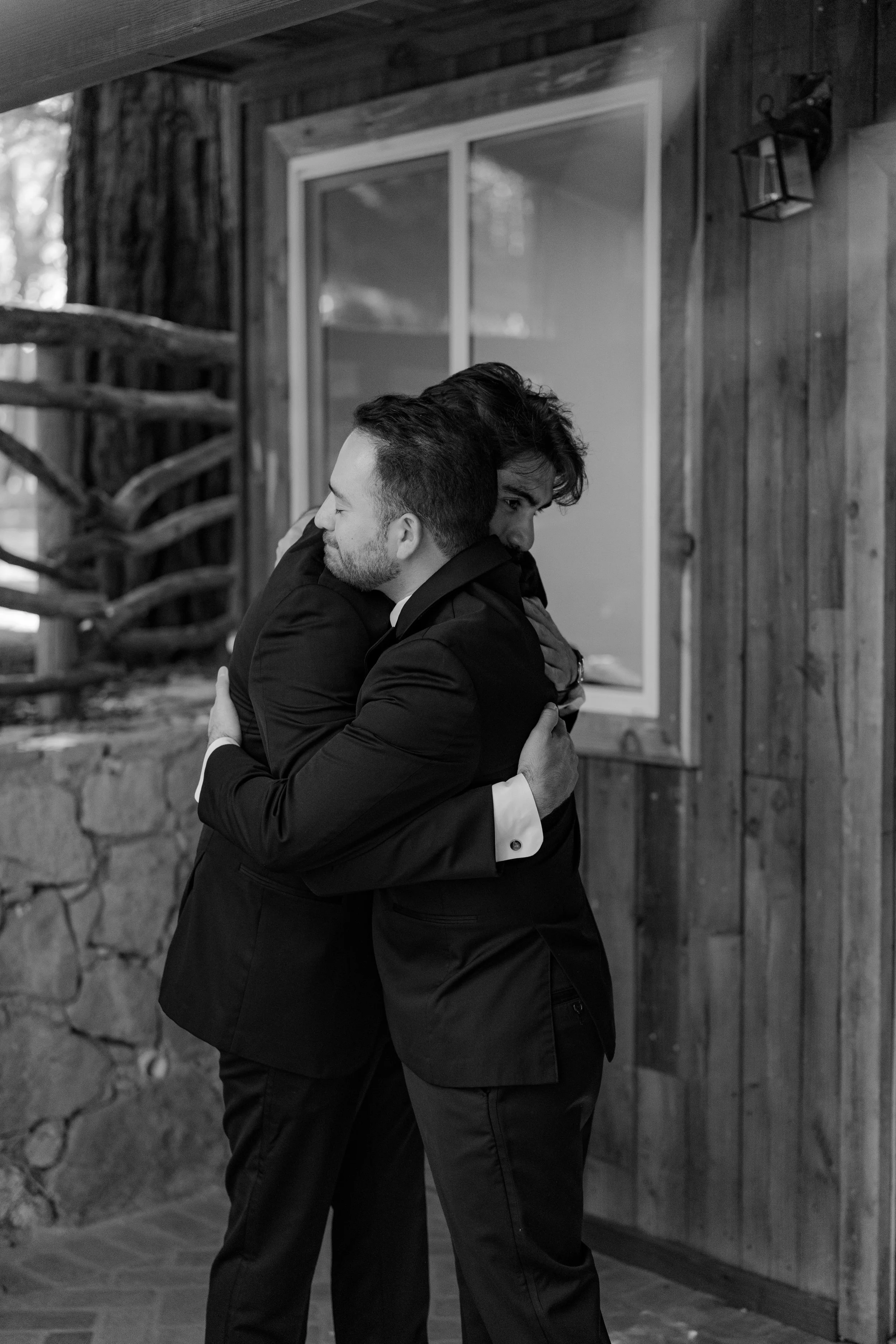 Two men in suits hugging each other indoors with wooden walls and large window in the background.