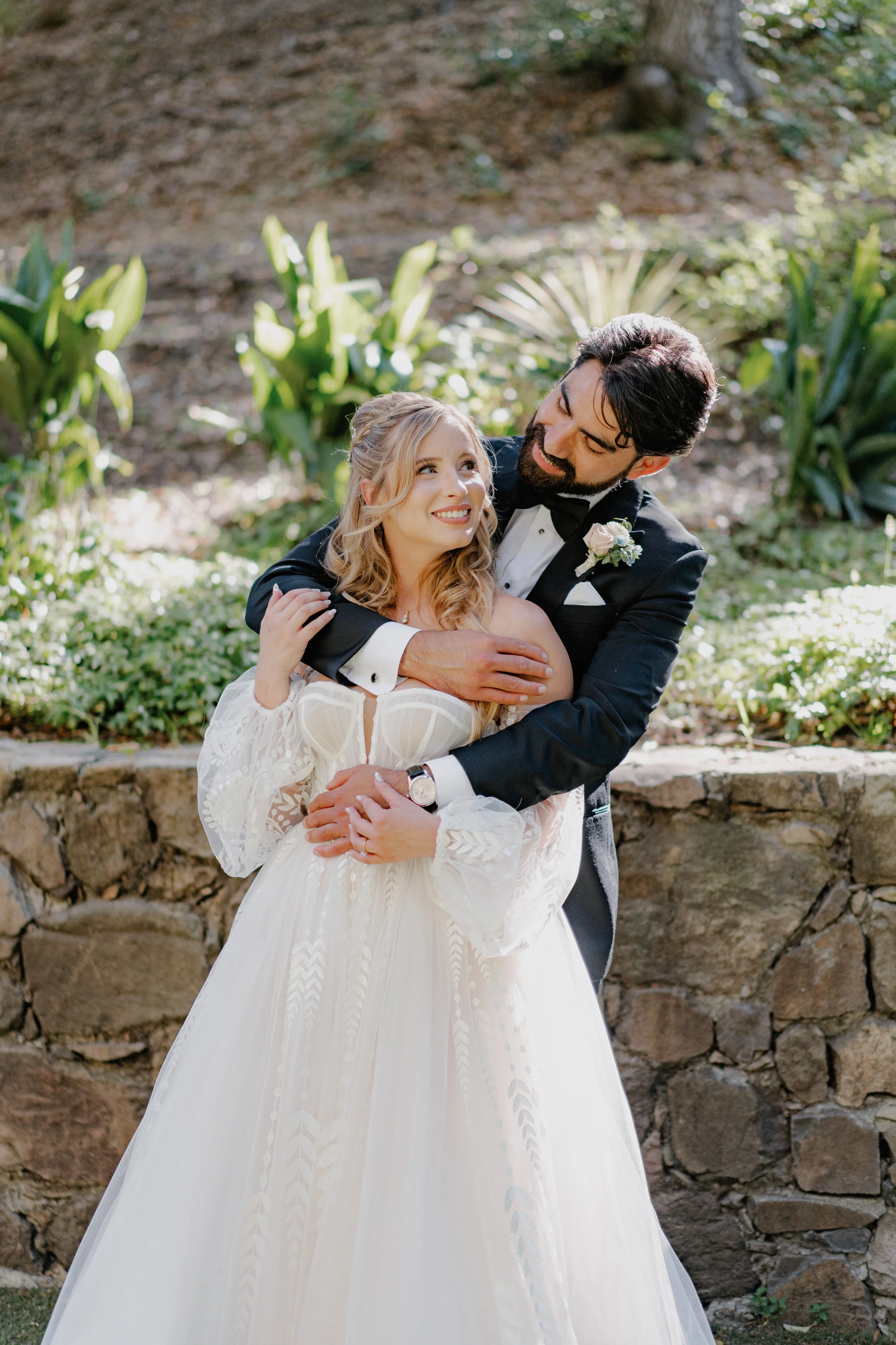 A bride and groom share a joyful moment outdoors, with the groom embracing the bride as they smile at each other. The bride wears a white wedding dress, and the groom is in a tuxedo with a boutonniere. Green plants and a stone wall are in the background.