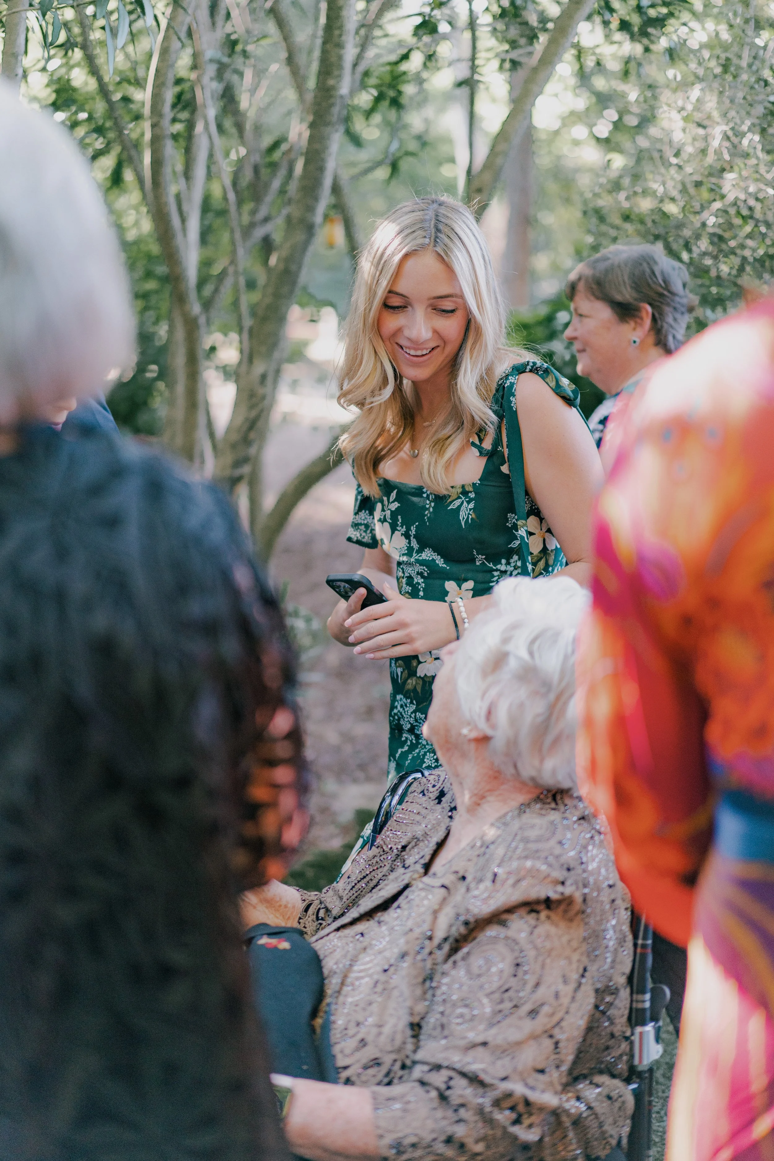 A young woman holding a phone and wearing a green floral dress is smiling and talking to an elderly woman sitting in a wheelchair outdoors surrounded by other women. The background is a garden with trees.