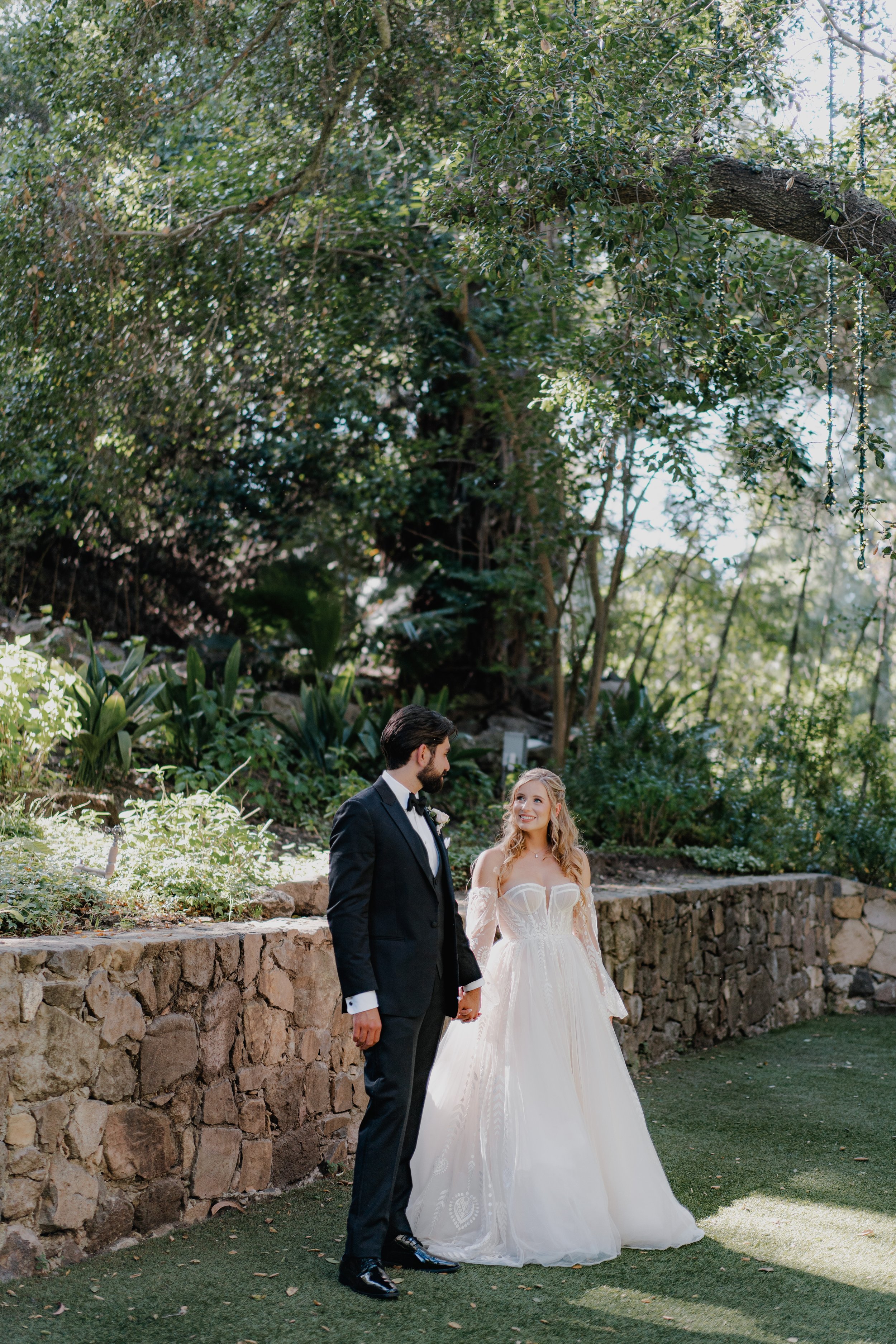 A bride and groom holding hands and standing outdoors in a garden, with tall trees and greenery in the background. The groom wears a black tuxedo and the bride wears a white wedding gown with off-the-shoulder sleeves.