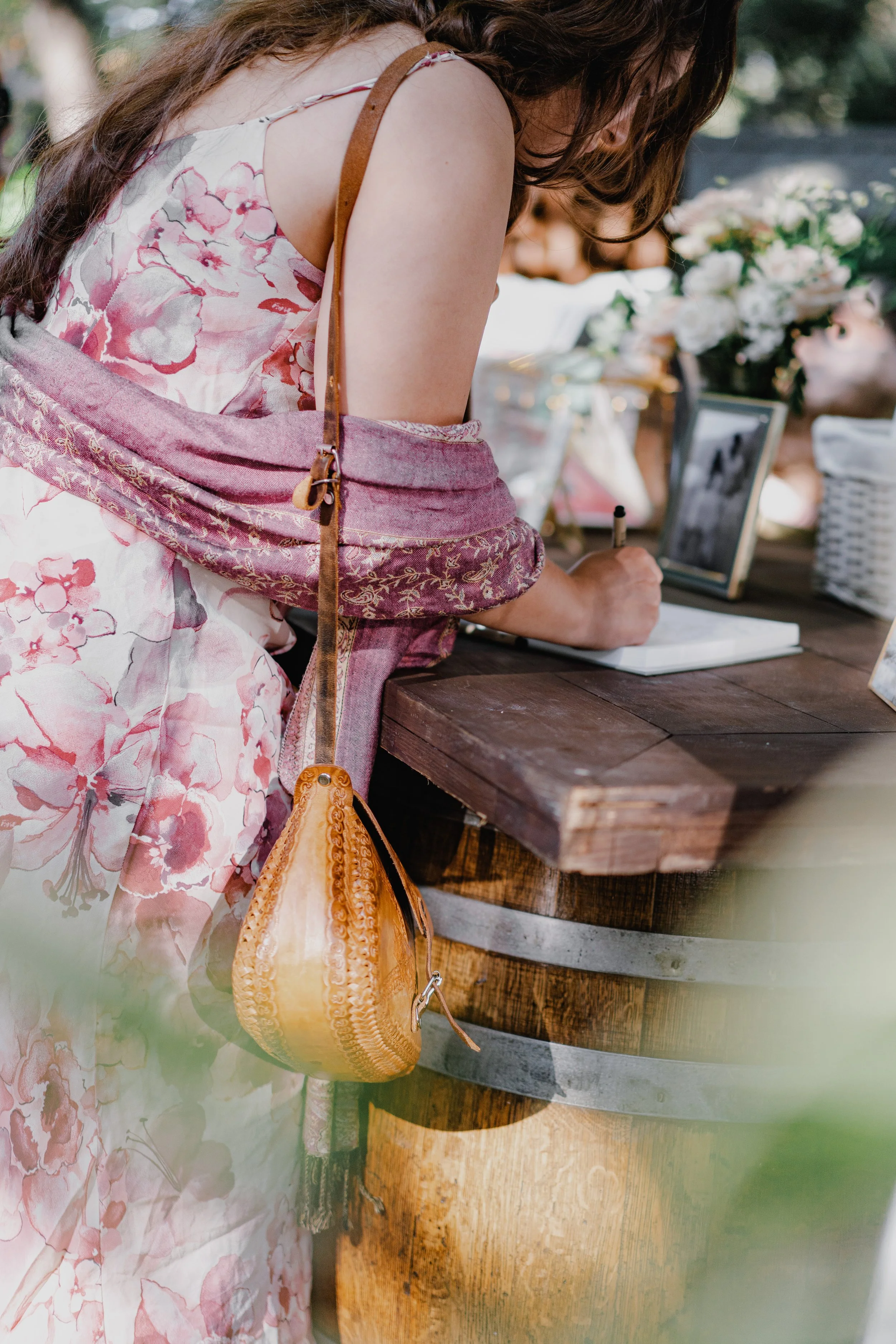 A woman in a floral dress signing a book at an outdoor event surrounded by flowers and framed photos.