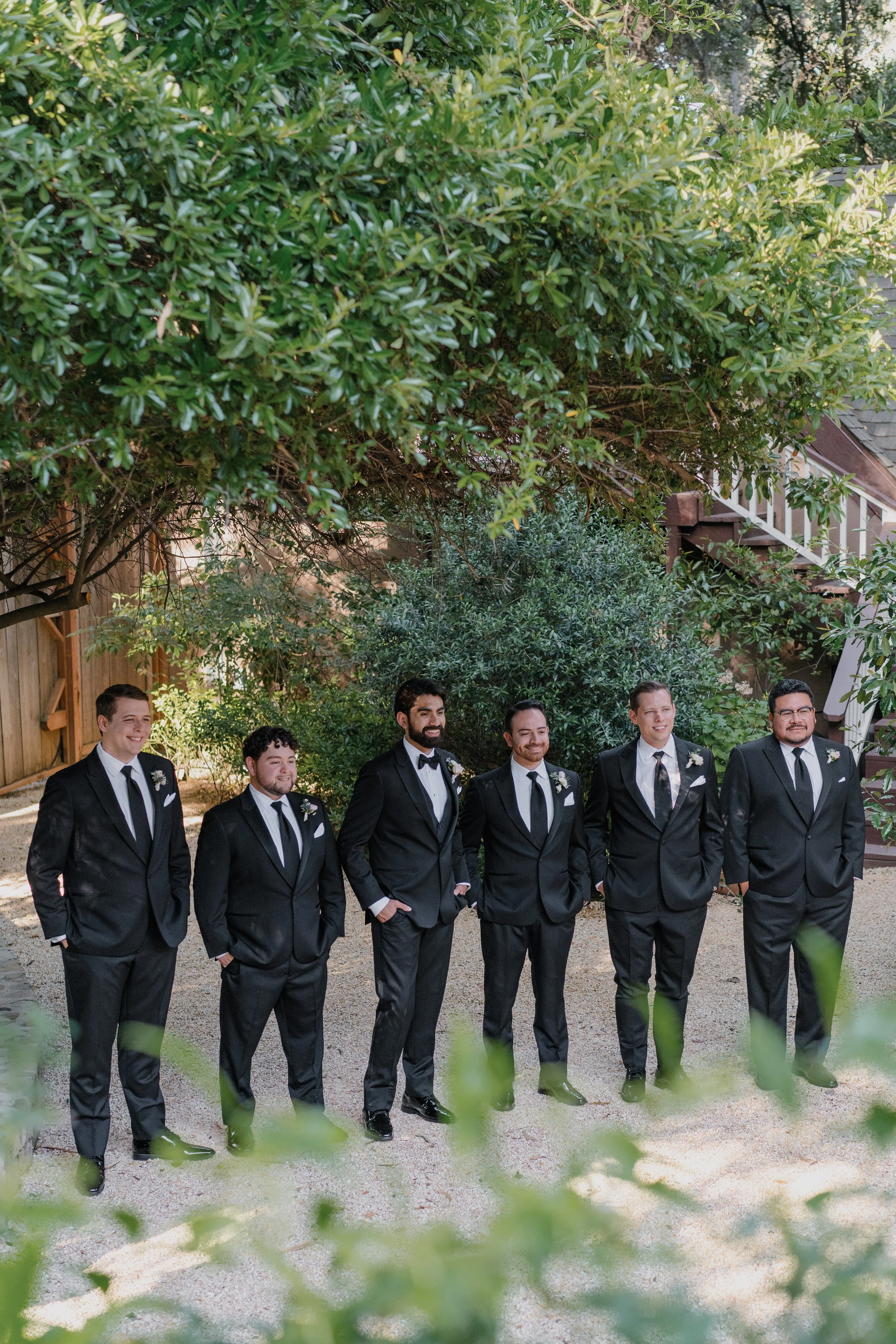 A group of seven men in tuxedos standing outdoors under a leafy tree, smiling. They are dressed for a formal event, likely a wedding, with white shirts, black ties, and boutonnières.