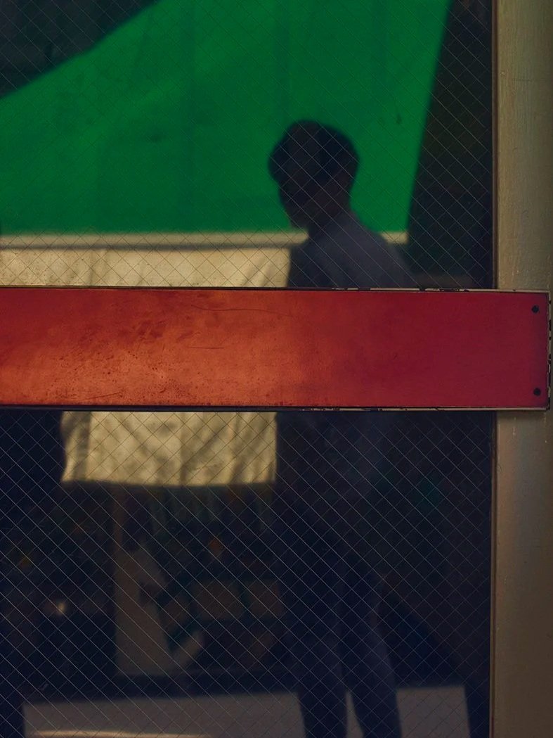 Reflet d'une personne derrière une vitre avec une fenêtre divisée en sections, une partie verte, une partie en or, et une partie rouge.