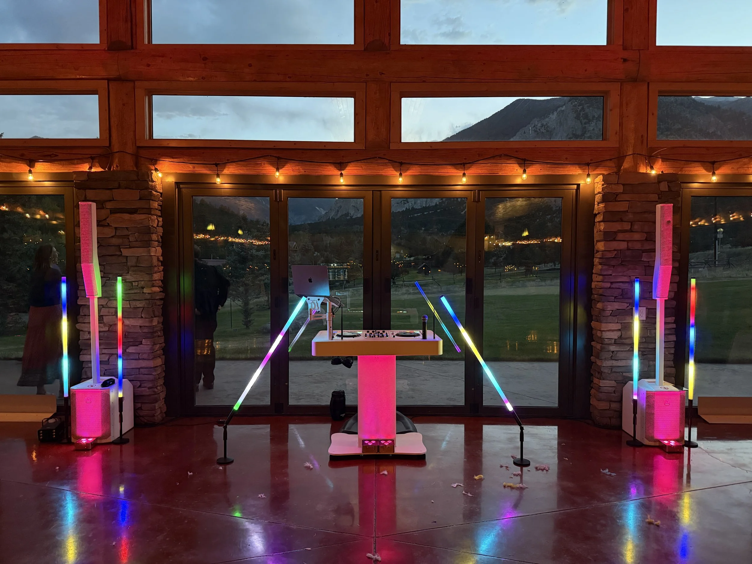 Indoor event space with colorful neon lights, a DJ setup, and large windows showing mountains and a landscape at dusk.