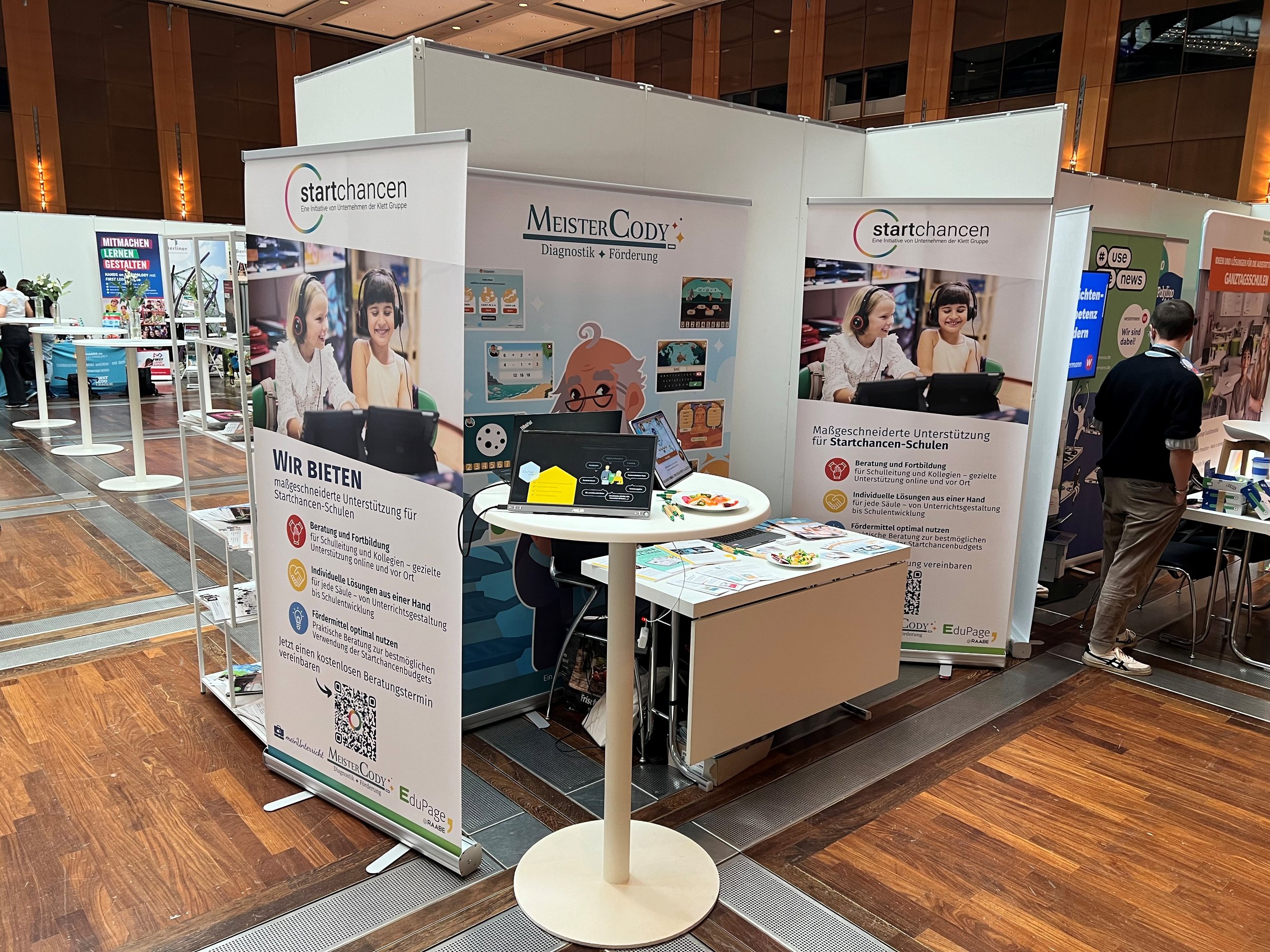 Messestand von Bildungschancen.schule auf dem DSLK 2024. Zu sehen sind Roll-ups von Startchancen und Meister Cody mit Informationen.
