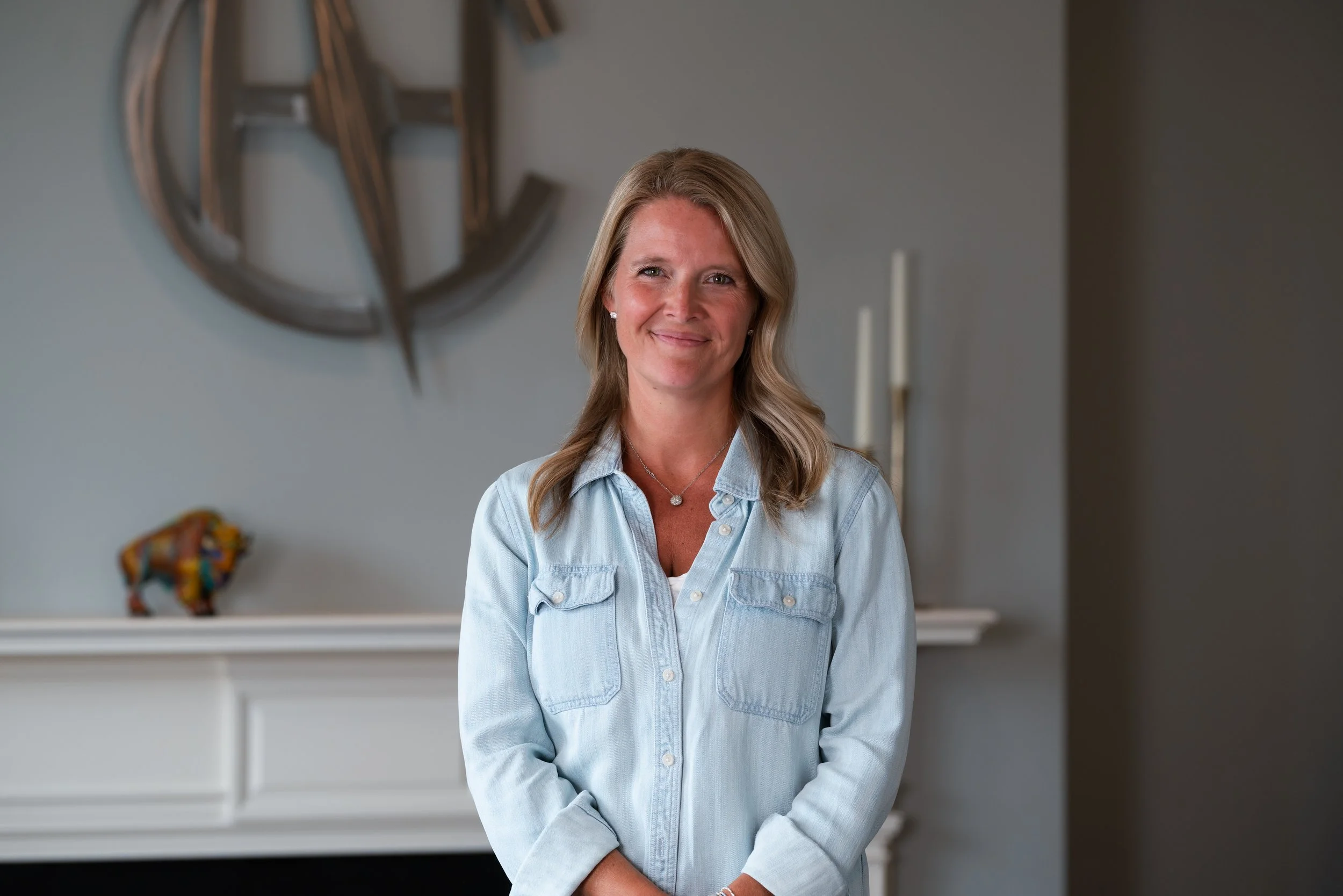 A woman with blonde hair, wearing glasses, a white T-shirt, olive green jacket, and jeans, standing near a window with natural light coming in.