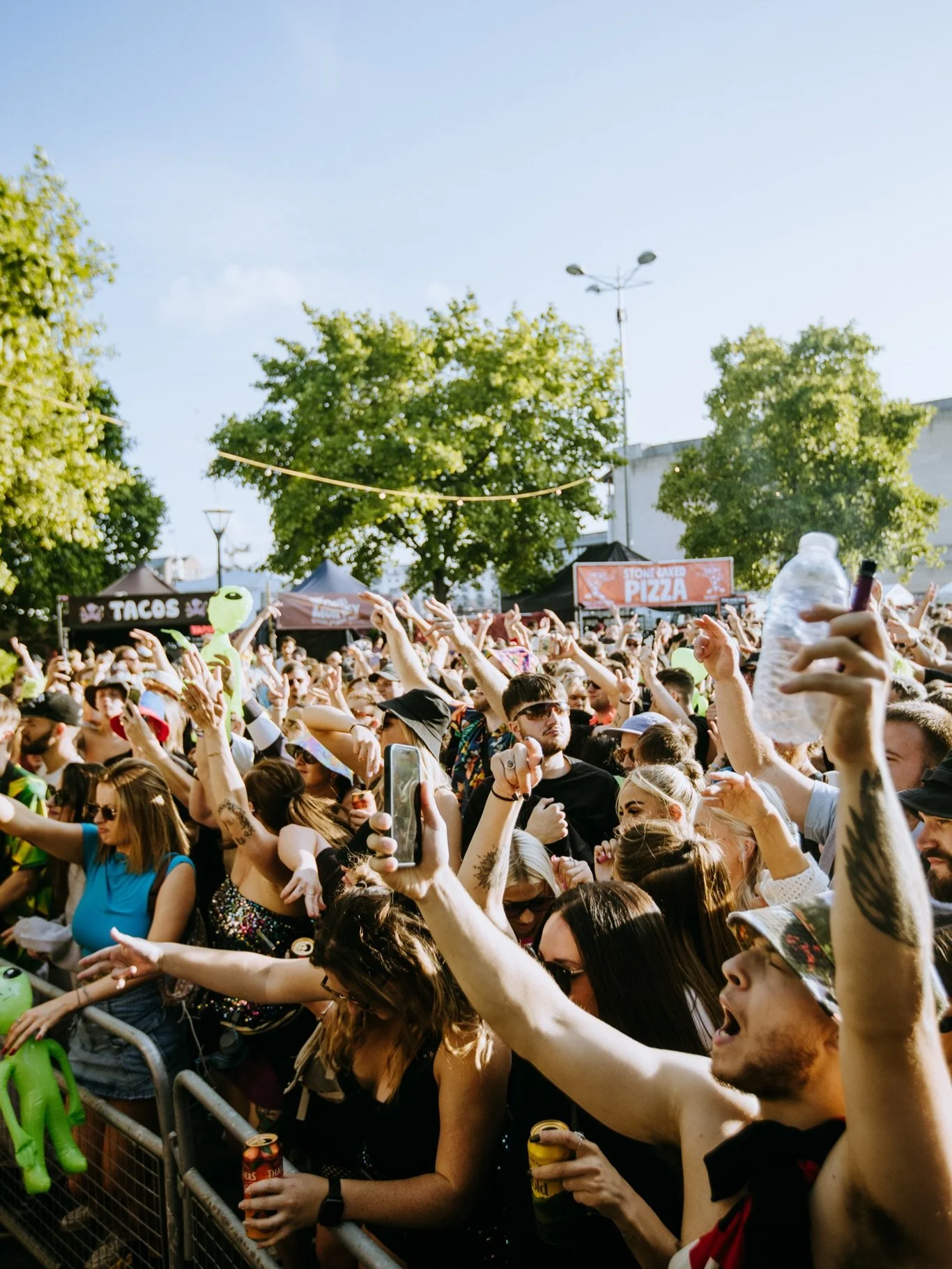 People enjoying a concert or festival, with two women sitting on men's shoulders, raising their hands and dancing in an indoor venue with brick walls.