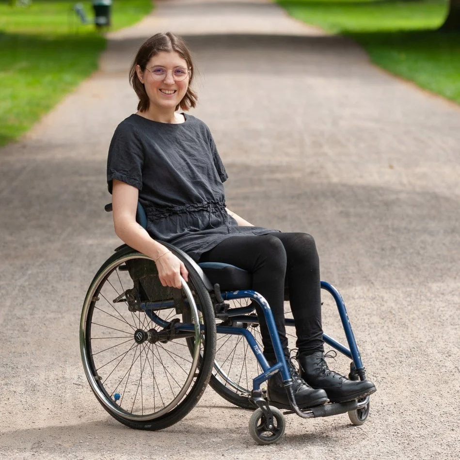 Bildbeschreibung: Mechthild sitzt auf ihrem Rollstuhl auf einem Kiesweg und lächelt in die Kamera.