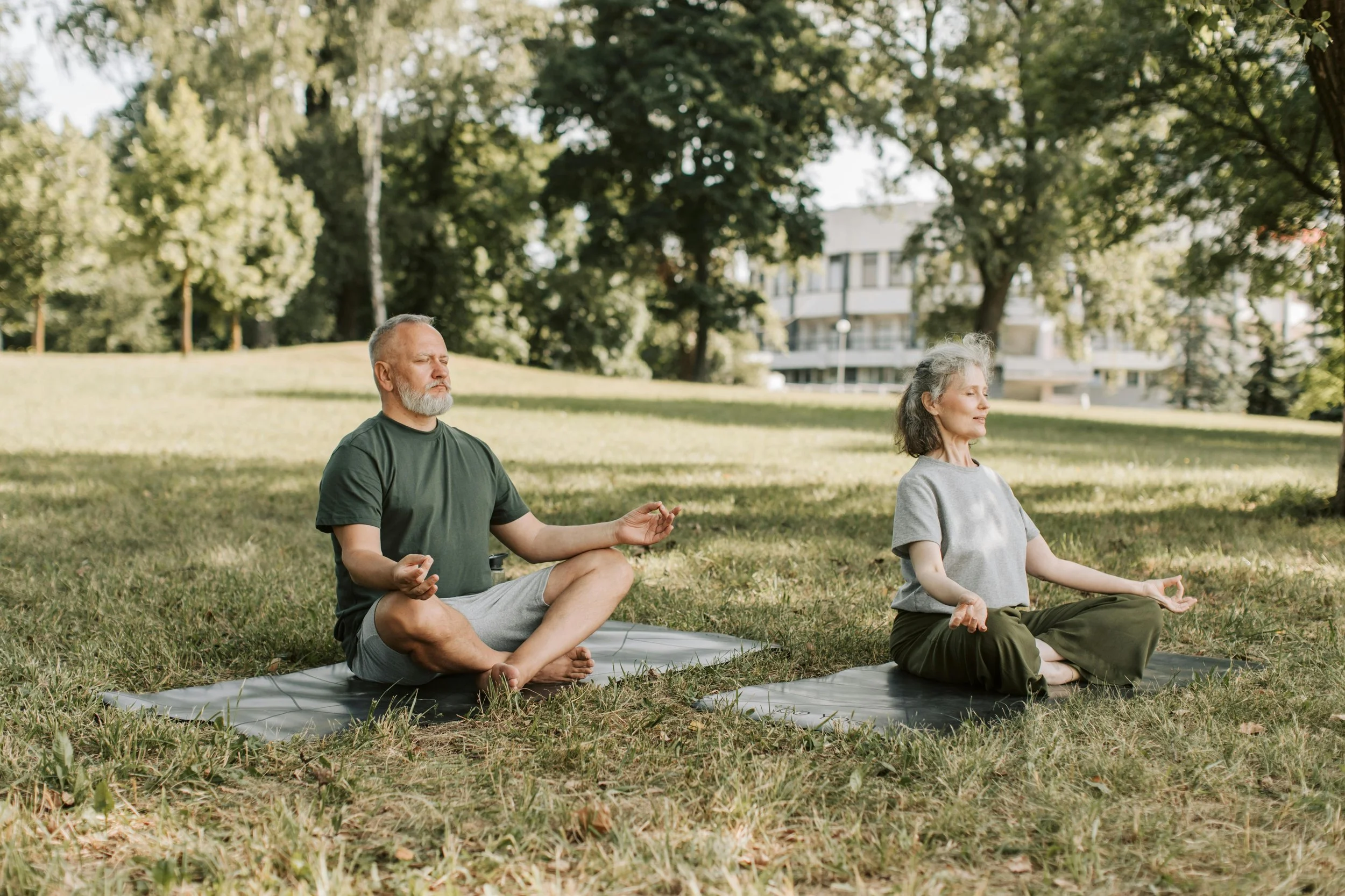 Zwei Personen auf Matten, die in Meditationshaltung auf einer grünen Wiese sitzen.