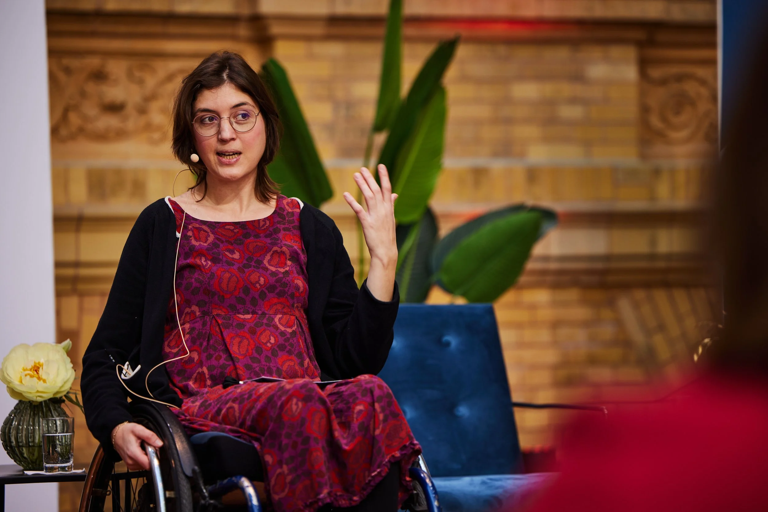 Mechthild sitzt im Rollstuhl auf einer Bühne und spricht, dabei gestikuliert sie mit den Händen. Im Vordergrund sieht man eine weitere Person von hinten und unscharf.