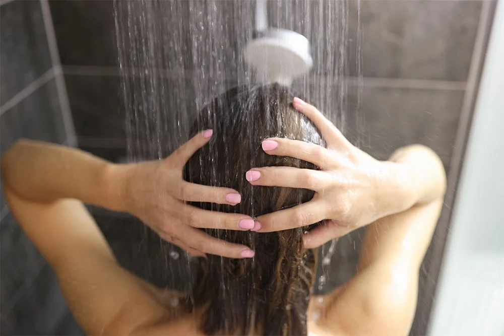 Person rinsing hair in shower with water streaming down in a bathroom shower stall.