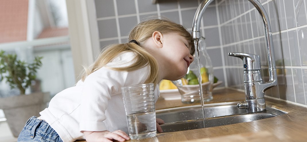 A young girl is leaning over a kitchen sink with her head under running water, appearing to enjoy washing her hair or playing in the water. She holds a glass of water in one hand.