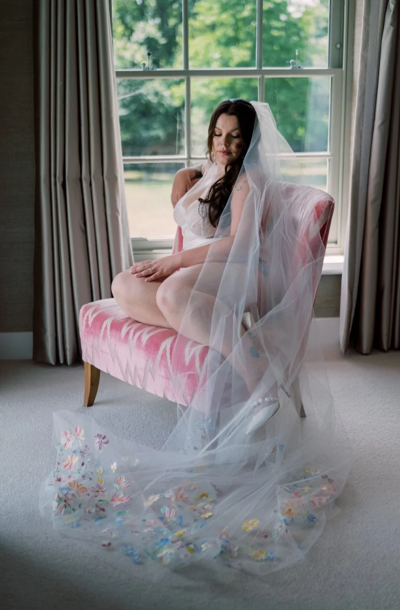A woman dressed in white lingerie with a wedding veil sitting on a pink velvet chair by a large window with green trees outside. A sheer veil with colorful floral embellishments is draped over her lap and the floor.