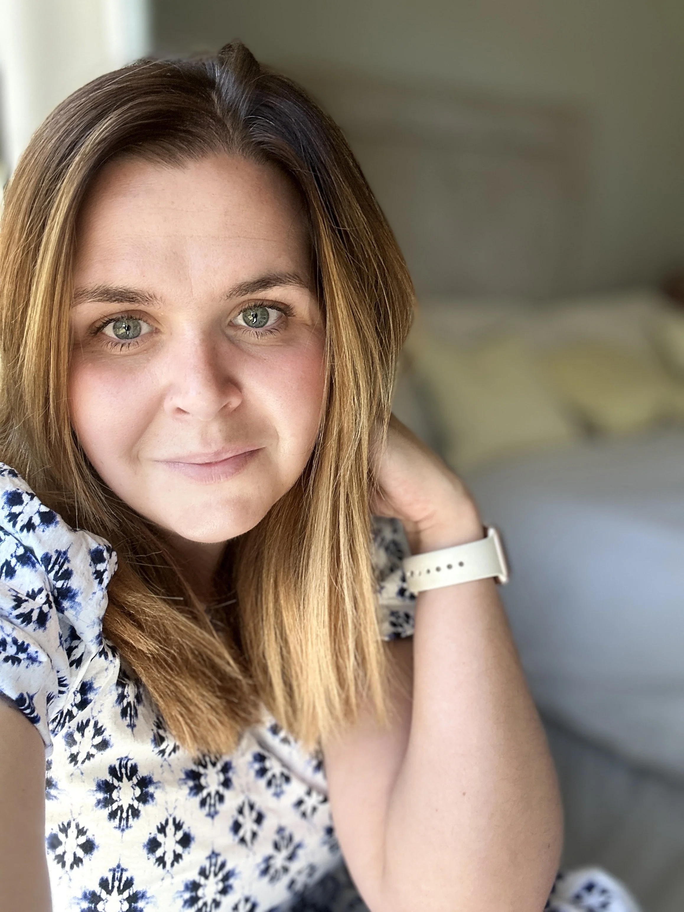 Close-up of a woman with shoulder-length light brown hair, blue-green eyes, and fair skin, smiling softly while sitting indoors near a window.