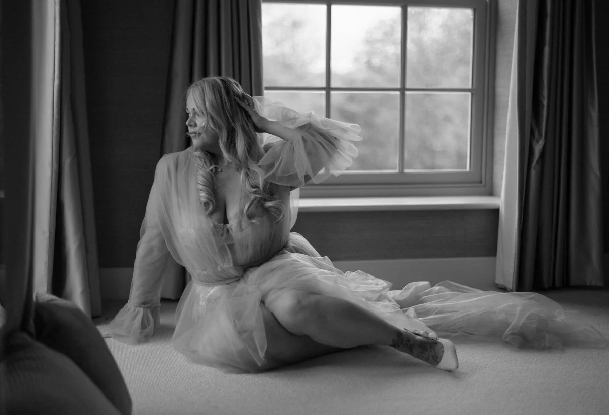 A woman with long hair sitting on the floor in front of a window, wearing a sheer, flowing dress, smiling and touching her hair.