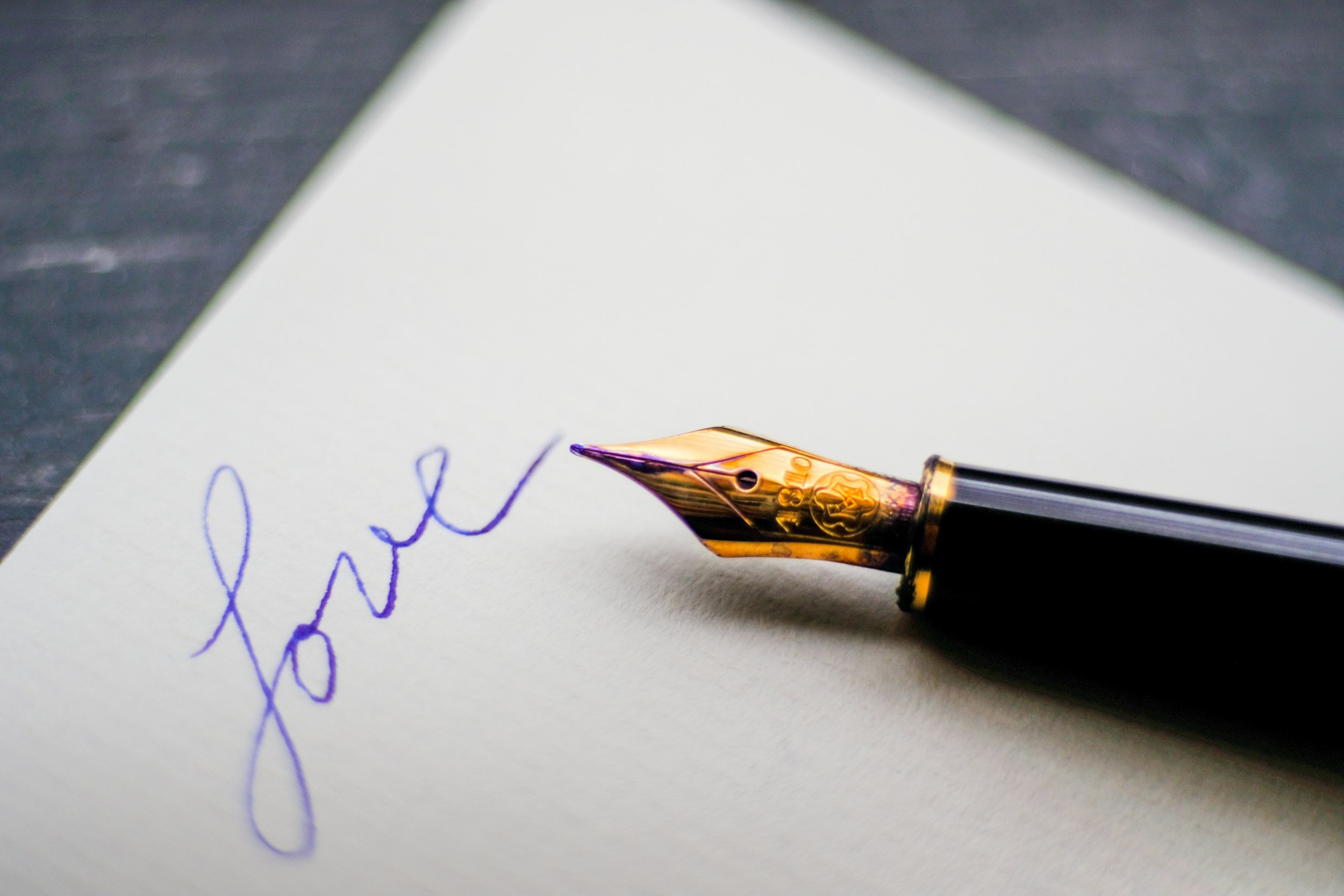 Close-up of a gold-tipped fountain pen writing on a white piece of paper. The handwriting in blue ink appears to spell out a word.
