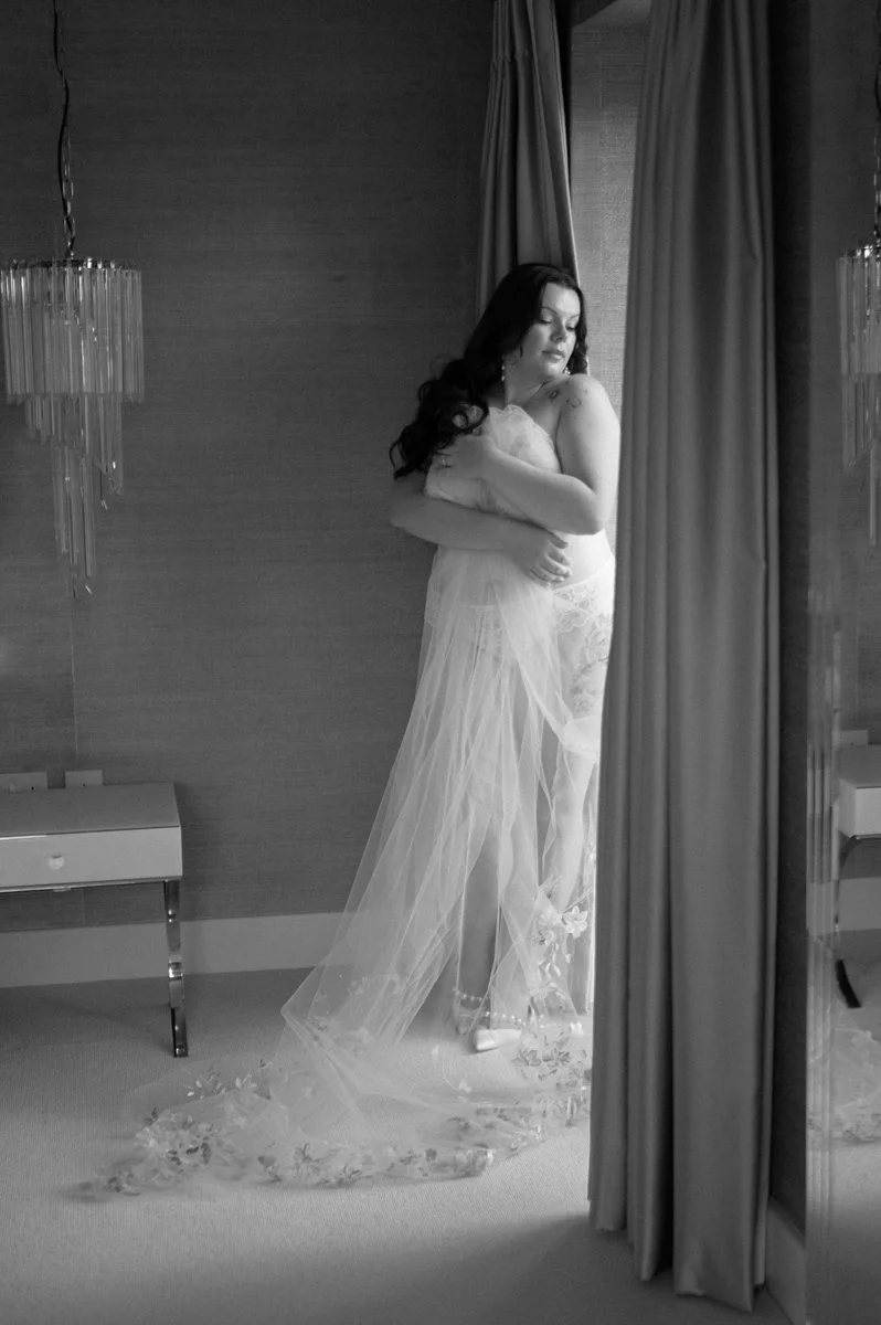 A woman in a sheer, white, lace dress standing in front of a curtain, looking down with her arms crossed over her chest.