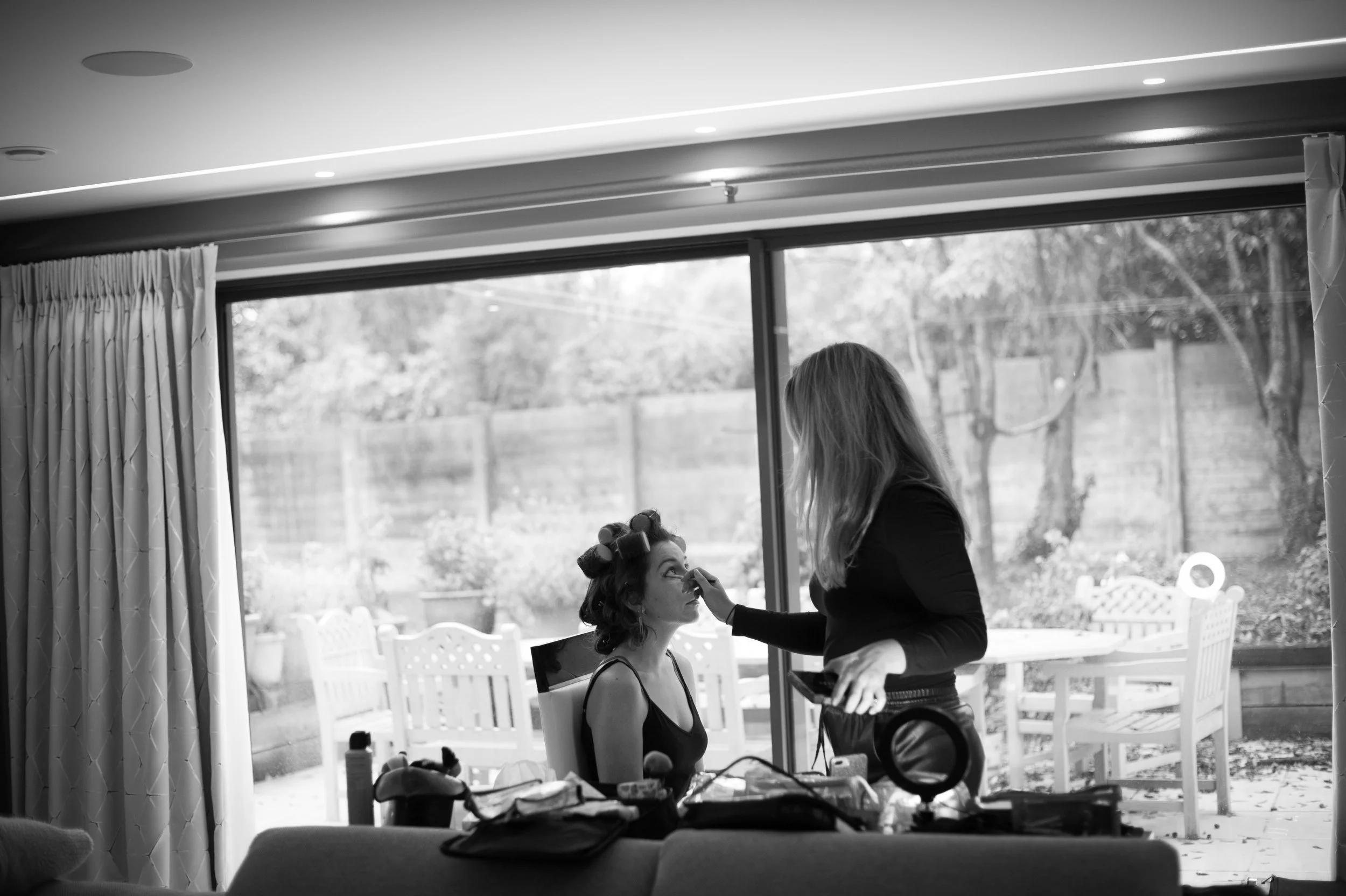A woman with rollers in her hair gets her makeup done by a makeup artist inside a room with a large window looking out to a backyard with trees and a wooden fence.