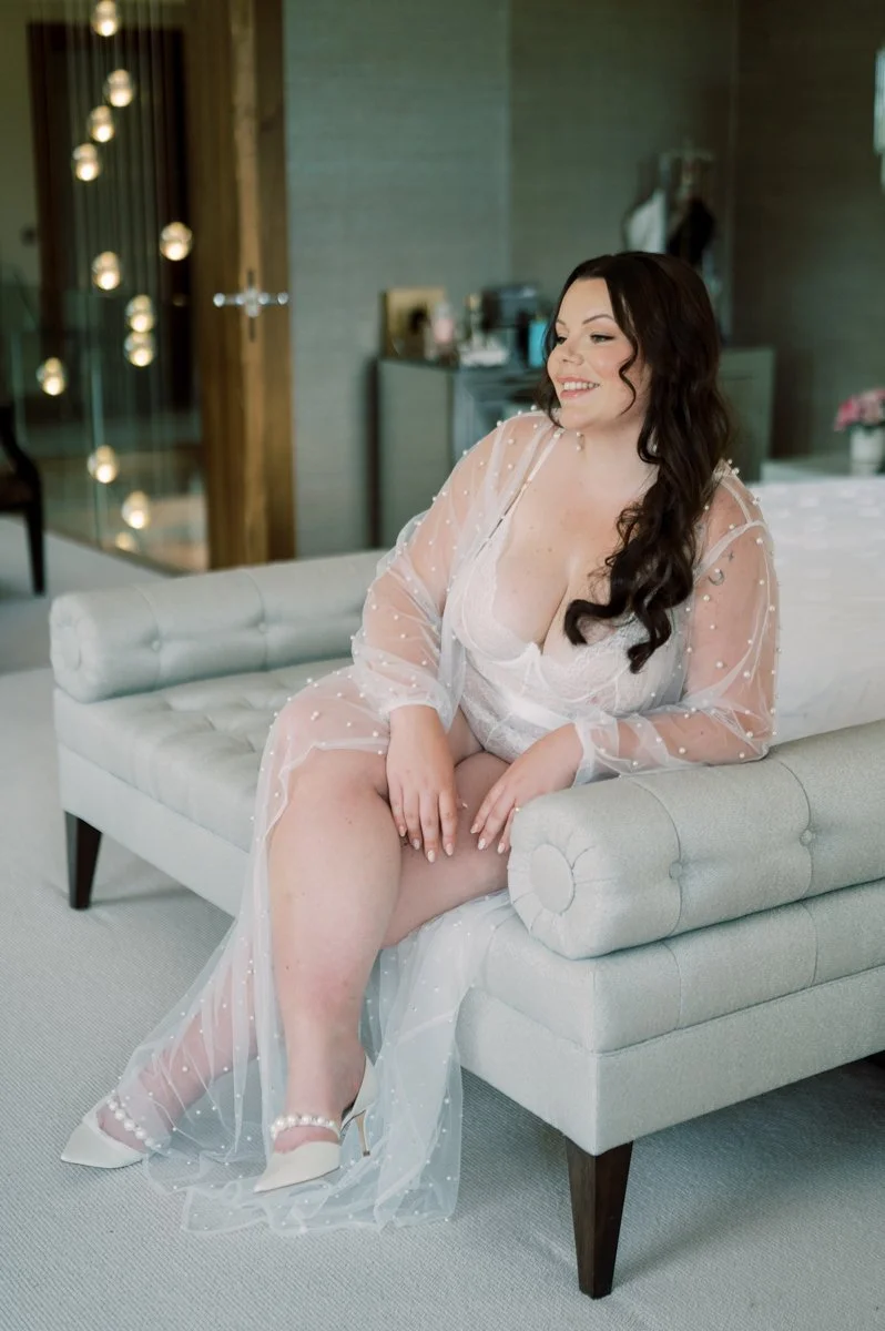 A woman with long dark hair in a white bridal robe sitting on a light-colored couch, smiling inside a modern room with soft lighting and decorative string lights.