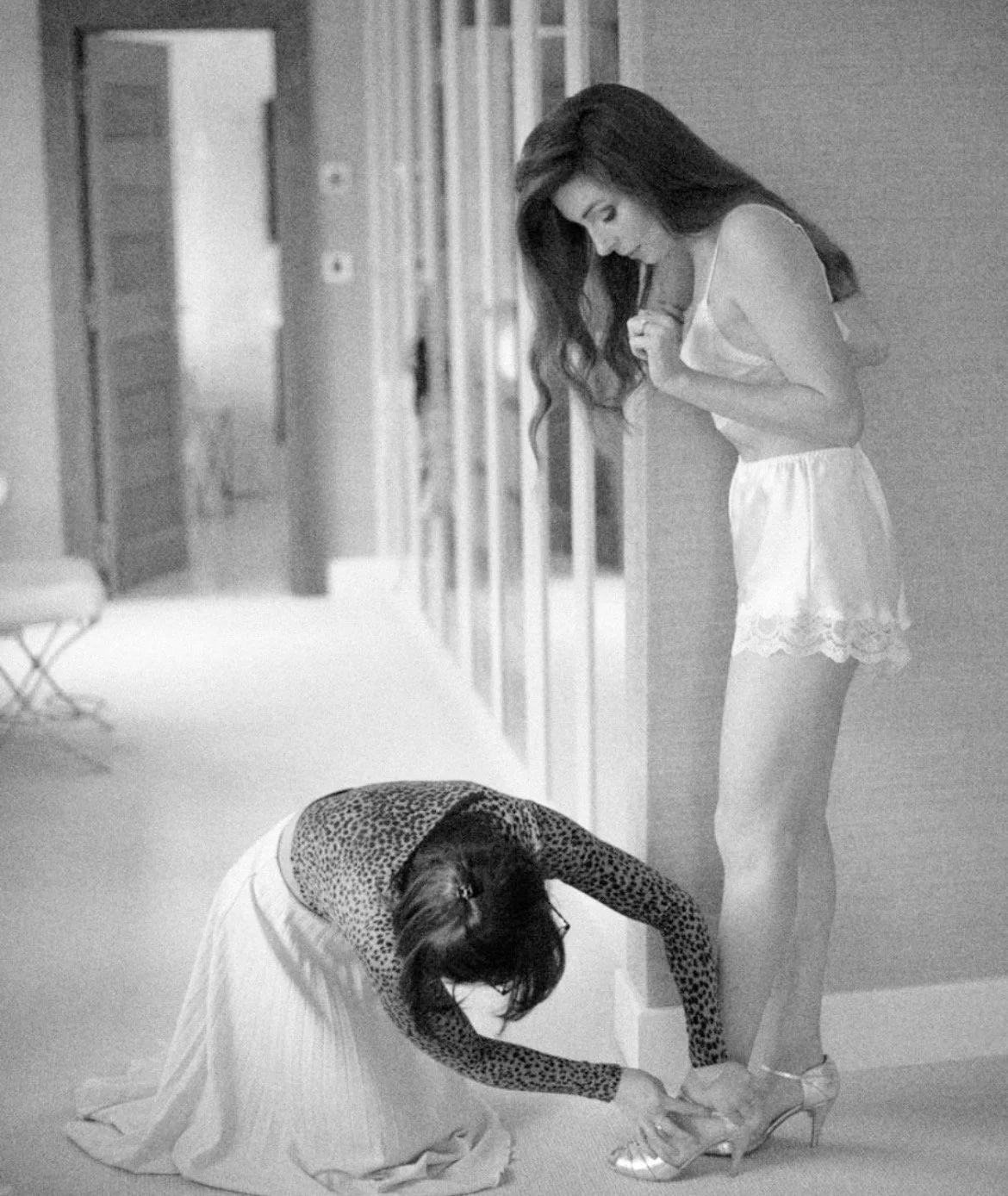 Two women in a hallway, one kneeling and helping the other with her shoes, both in nightwear.