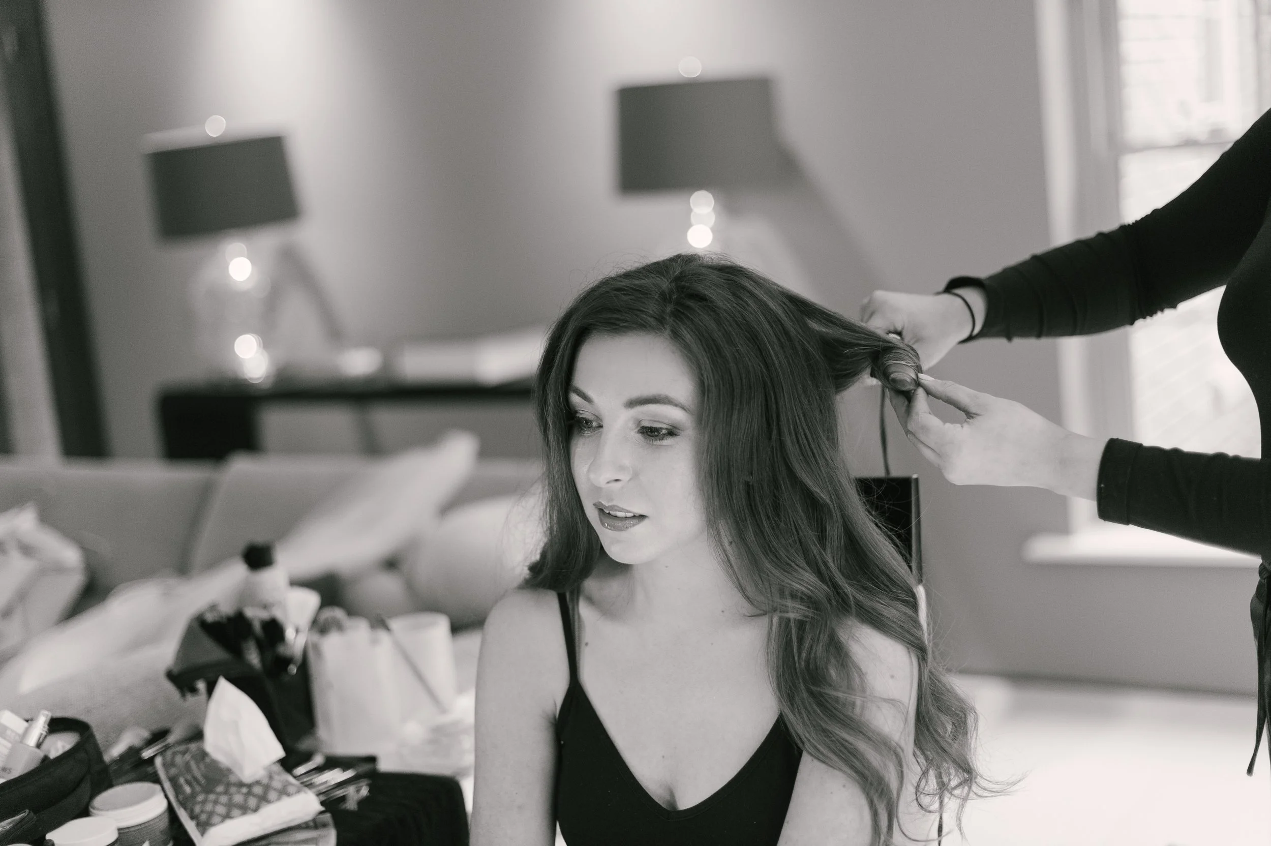 A woman with long wavy hair sitting on a sofa as a hairstylist styles her hair in a bright room with lamps and a window.