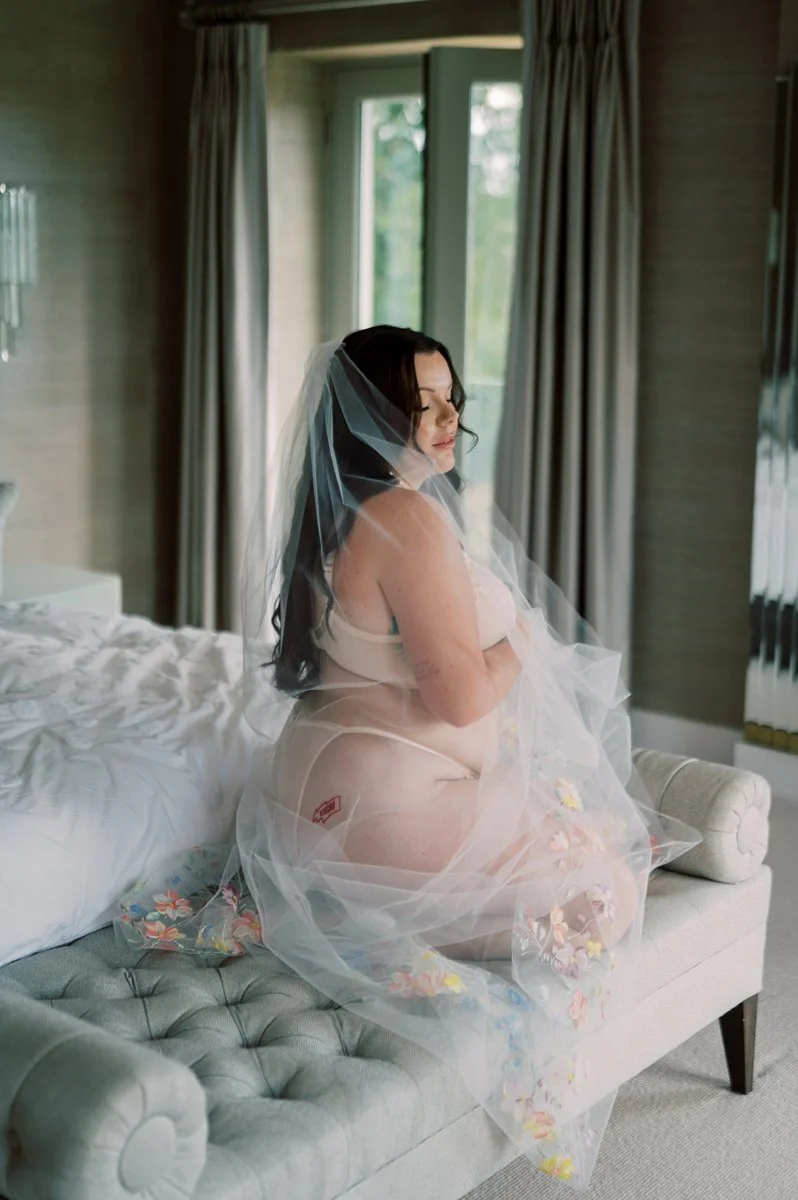A woman with long dark hair and light skin, wearing nude-colored lingerie and a bridal veil, sitting sideways on an upholstered bench in a softly lit room near a window with green foliage outside.