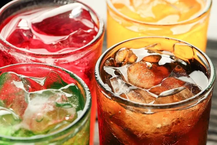 Four colorful glasses filled with different sodas and ice, including red, yellow, green, and dark brown beverages.