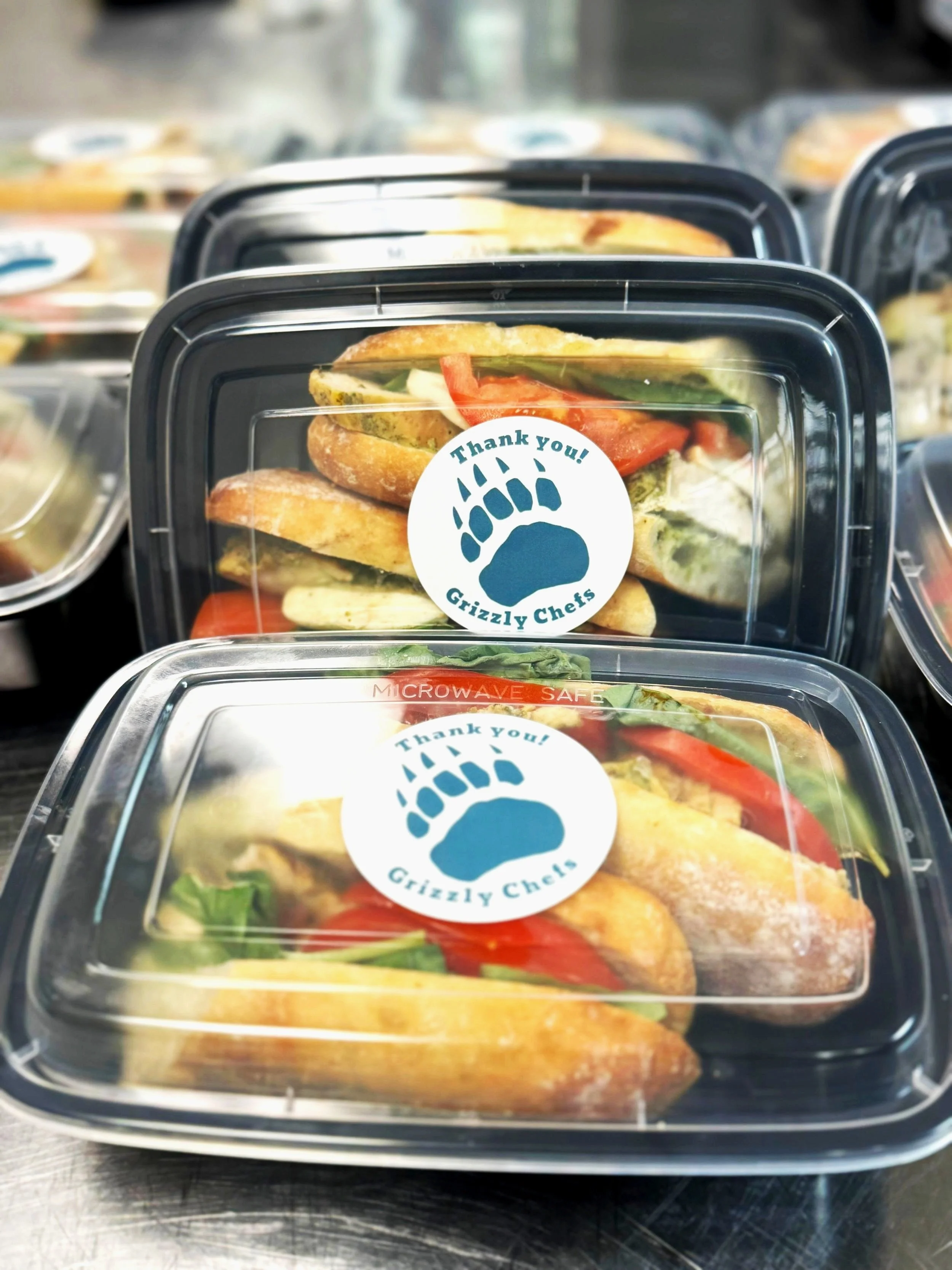 Pre-packaged sandwiches in clear plastic containers with a sticker that says "Thank you! Grizzly Chefs" and features a bear paw print.