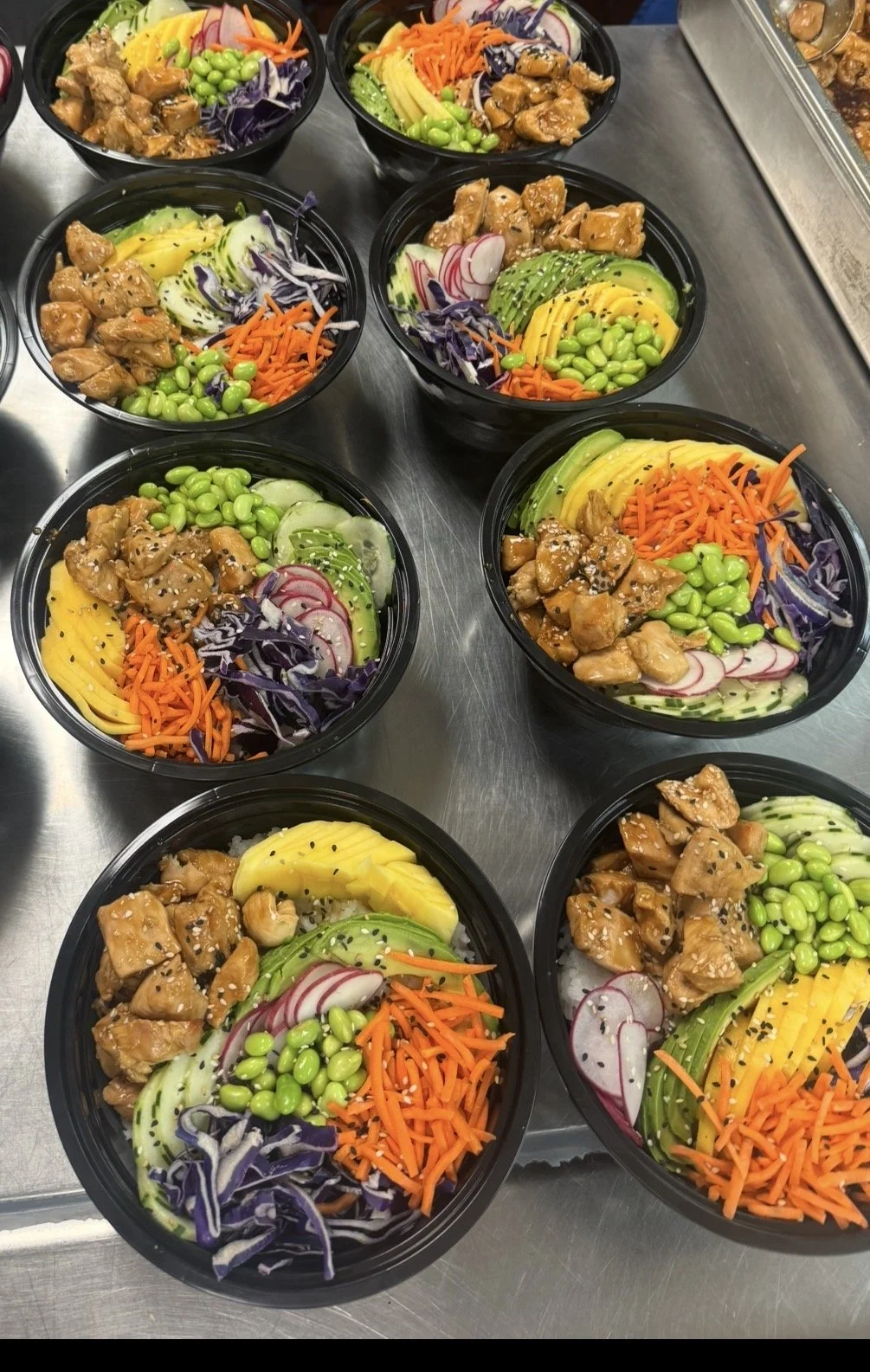 Multiple bowls of poke salad with diced chicken, sliced radishes, shredded purple cabbage, sliced cucumber, shredded carrots, sliced avocado, and mango on a stainless steel surface.