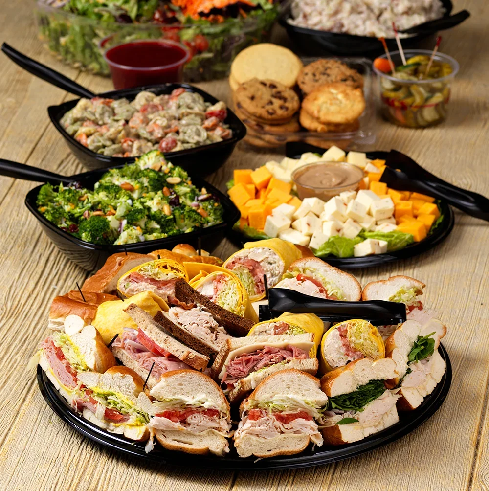 Assorted sandwiches with various meats and cheeses on a black platter, surrounded by salads, cookies, dips, and other side dishes on a wooden table.
