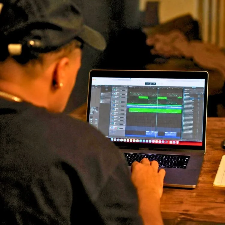 Independent music producer viewed from behind editing tracks on a laptop at a wooden table.