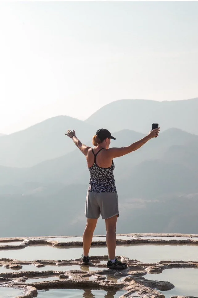 woman standing on plateau overlooking mountains with hands raised in the air holding cell phone