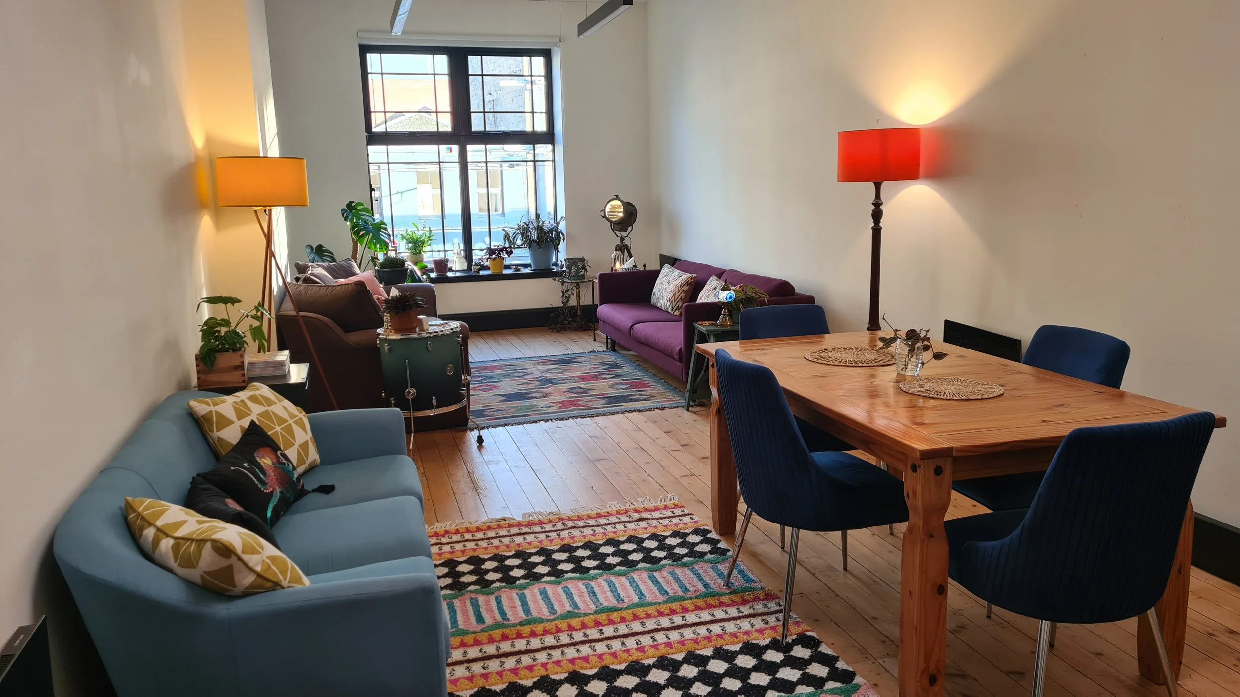 A cozy living room with a variety of colorful furniture, including a light blue sofa with patterned pillows, a purple couch, and a wooden dining table with blue chairs. The room has wooden floors, colorful rugs, and a large window with plants on the sill, letting in natural light. There are two tall floor lamps providing warm light, and a painting or textured wall on the left side.