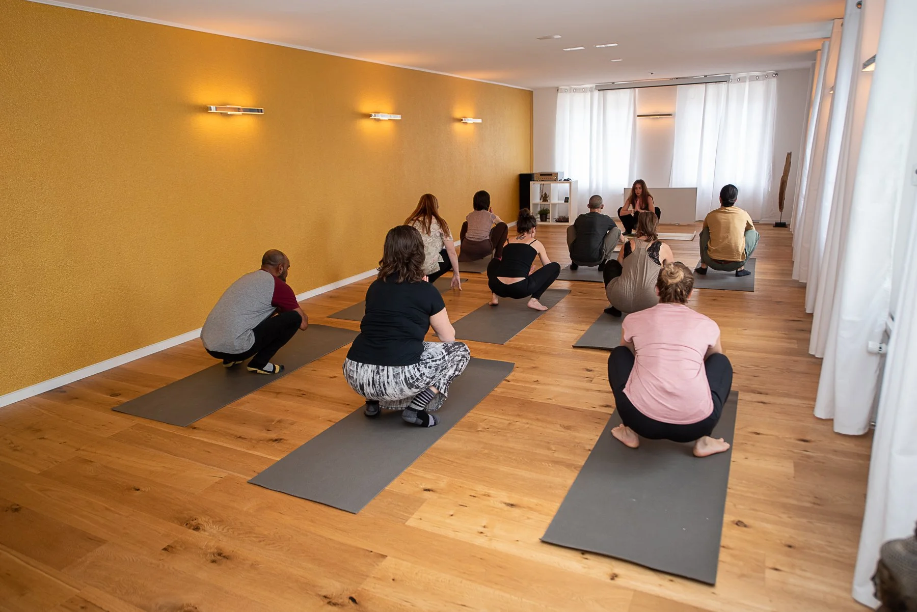  Eine Trainerin leitet eine diverse Gruppe von Menschen an, die in einem hellen Studio mit gelber Wand und Holzboden eine tiefe Kniebeuge-Pose auf Yogamatten ausführen. 