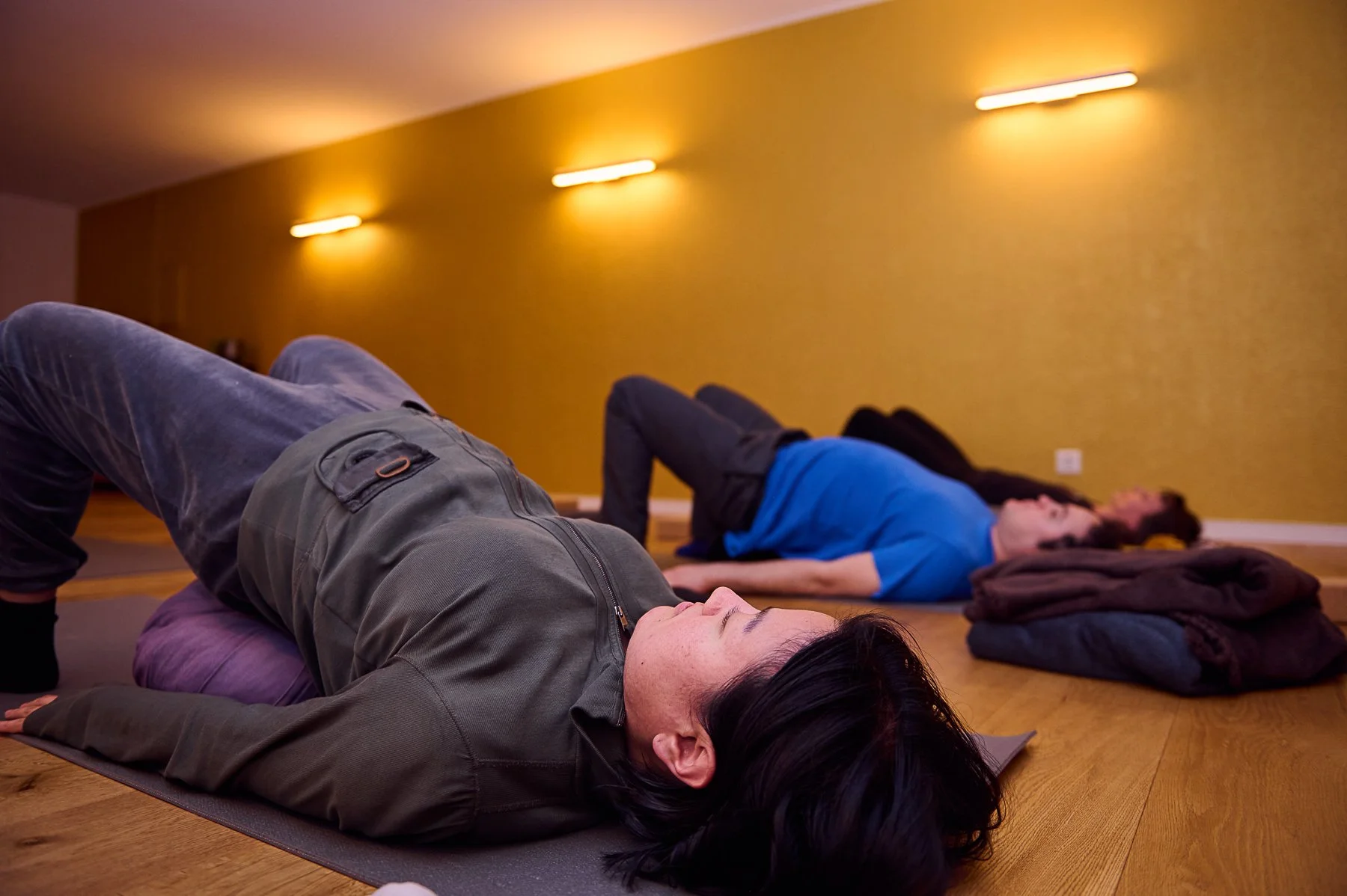  Teilnehmer liegen auf Yogamatten und führen eine unterstützte Schulterbrücke in einem gedimmten Studio mit gelber Wand aus, wobei Hilfsmittel wie ein lila Kissen unter dem unteren Rücken verwendet werden. 