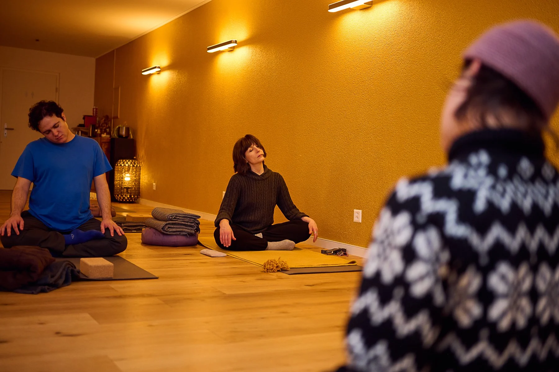  Zwei Teilnehmer sitzen im Kreuzsitz auf Yogamatten und dehnen ihren Nacken in einem gedimmten Studio mit gelber Wand, gesehen aus der Perspektive hinter einer Trainerin. 