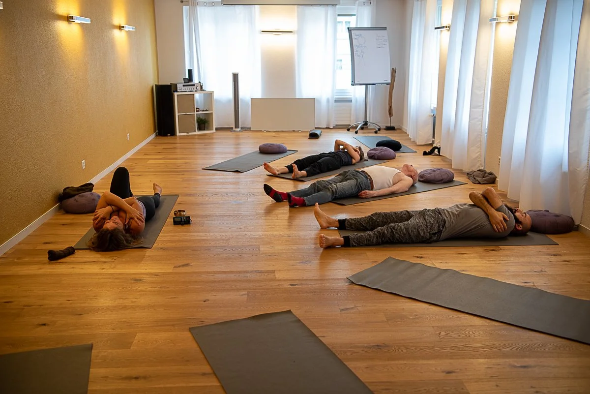  Mehrere Teilnehmer liegen auf Yogamatten in einem hellen Studio mit Holzboden; einige ruhen in der Totenstellung (Savasana), während andere während eines Kurses die Arme über der Brust kreuzen. 