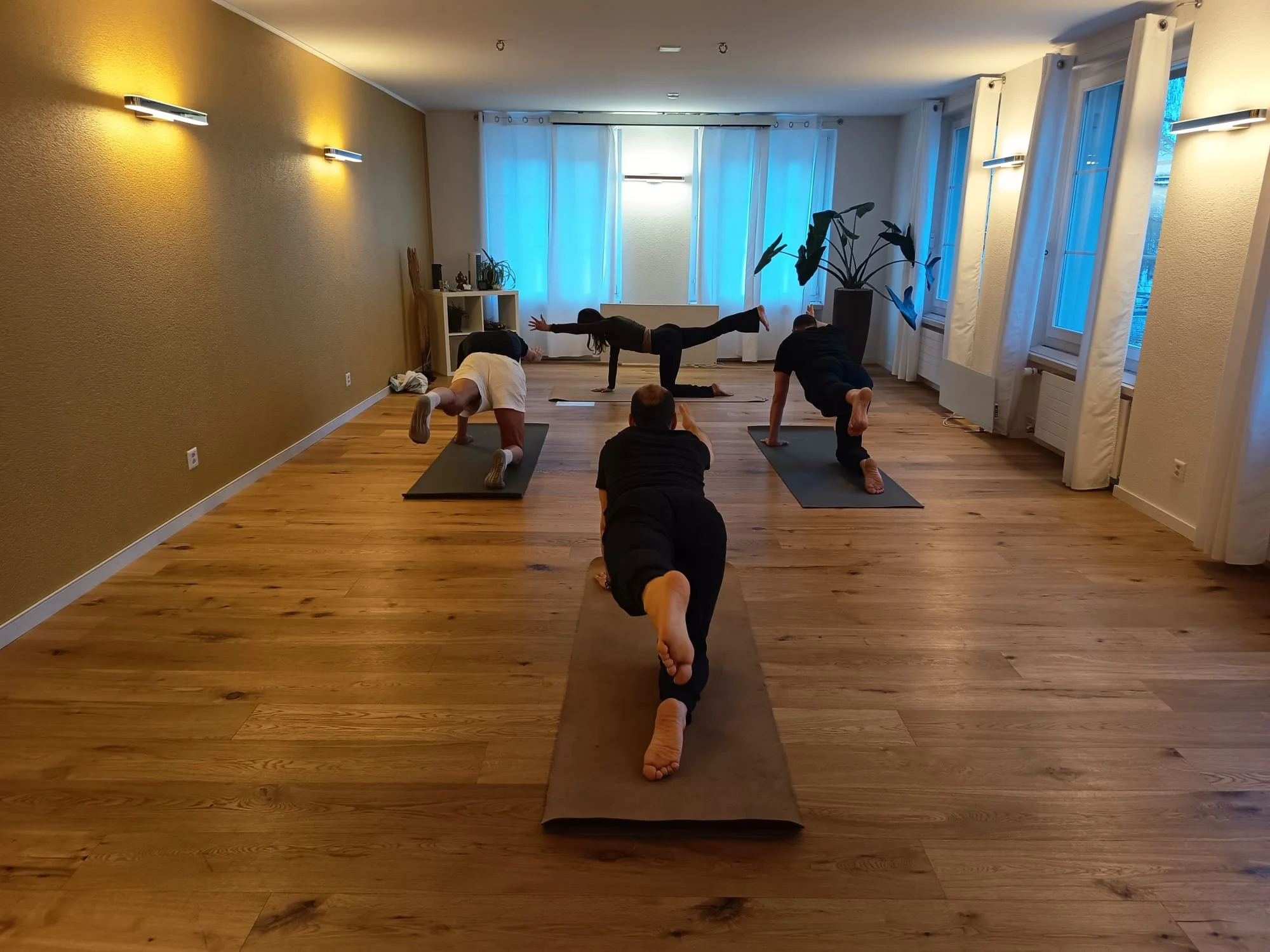  Eine Trainerin leitet eine kleine Gruppe von Teilnehmern an, die die Bird-Dog-Pose auf Yogamatten in einem geräumigen Studio mit Holzboden und einer gelben Akzentwand ausführen. 