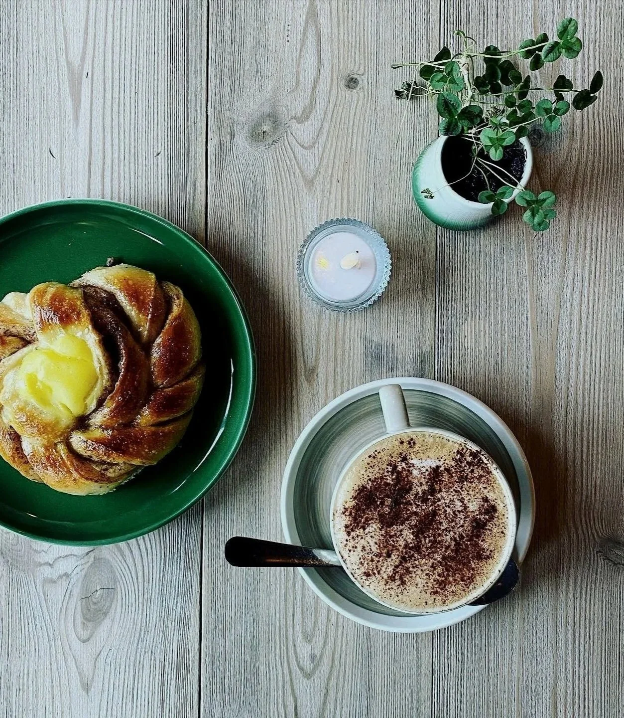 En kopp med kaffelatte, en bolle med kanel og melk, et grønt fat med en gjærbakst, en liten liten lys lysestake, og en liten blomsterpotte med grønn plante på et lyst trebord.
