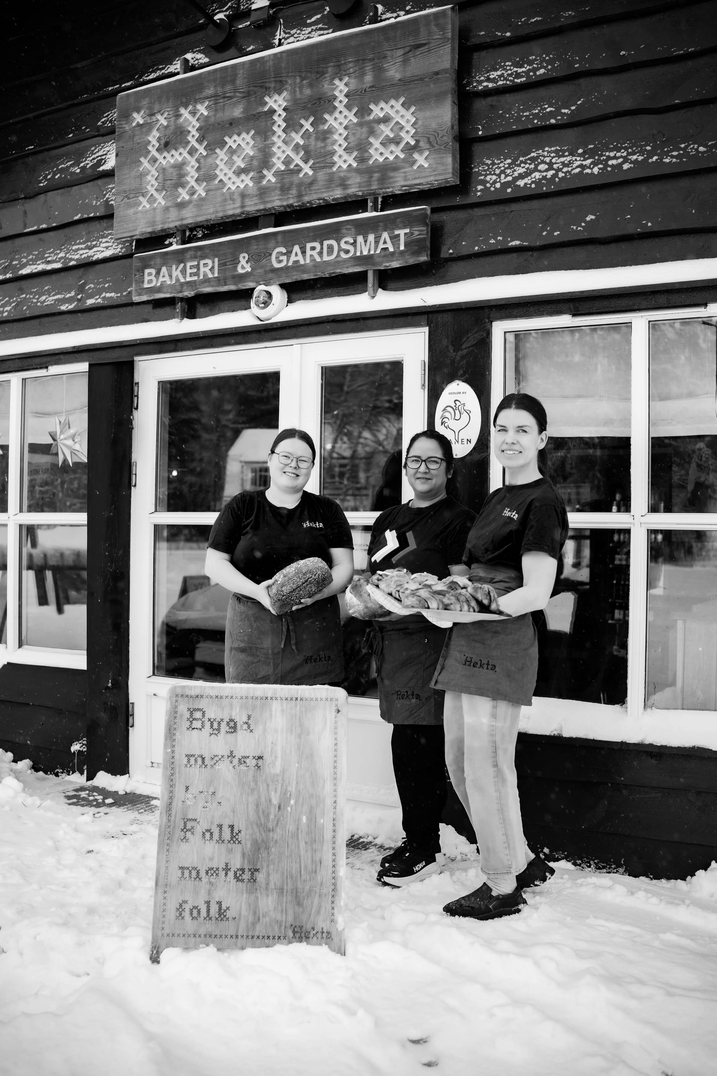Tre kvinner står utendørs i snø foran en bygning med tre vinduer. Alle har på seg svarte T-skjorter og holder brød og bakverk. Det er snø på bakken og på bygningens yttervegg, og det henger en dekorasjon i vinduet. Det er et skilt foran som sier 'Bygd baker' og har broderte ord.