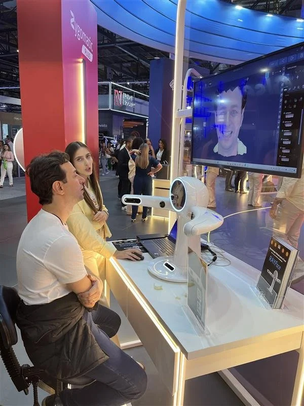 People interacting with a robotic AI assistant at a technology exhibition, with a large screen displaying a smiling face and a camera on a stand.