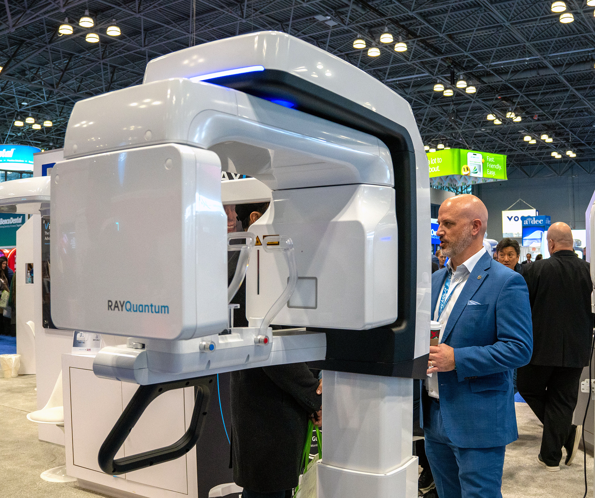 A man in a blue suit standing next to a large, white, advanced medical imaging machine labeled 'RAYQuantum' at a trade show or medical exhibition.