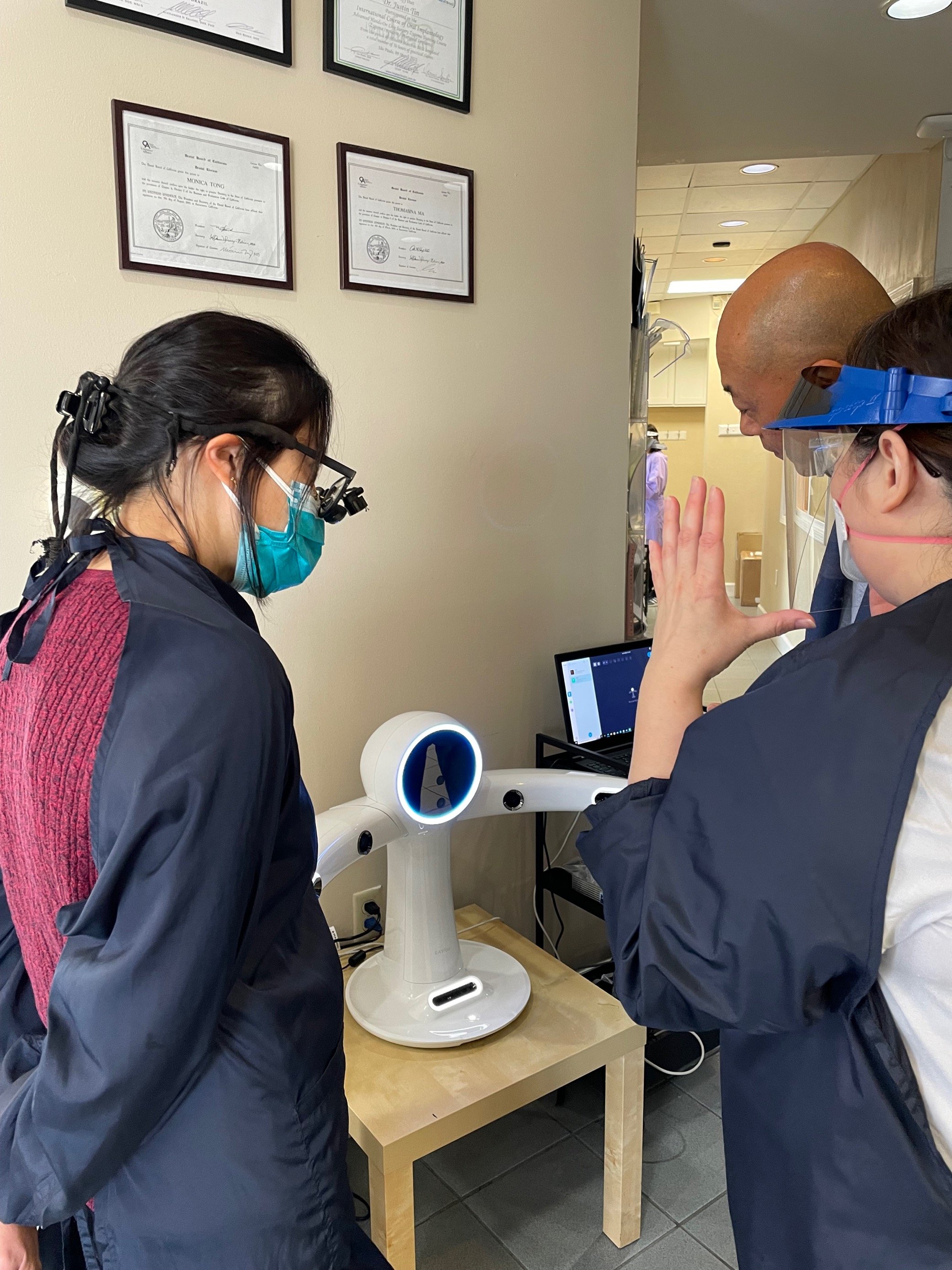 Medical professionals, wearing masks and protective gear, interact with a robot in a healthcare setting, with framed certificates or diplomas on the wall in the background.
