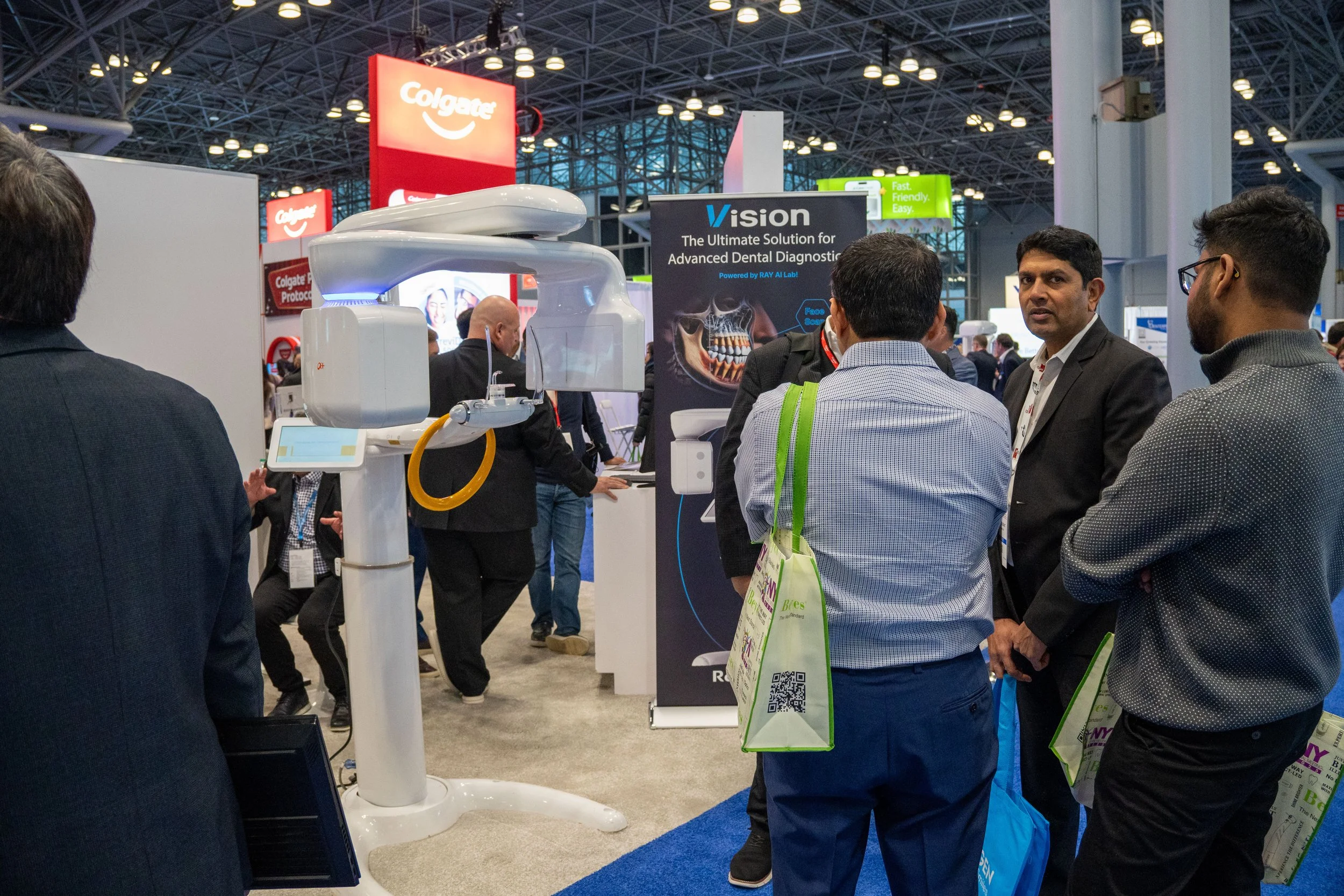 A group of people gathered around a large dental imaging machine at a trade show or exhibition, with a banner advertising advanced dental diagnostic technology in the background.
