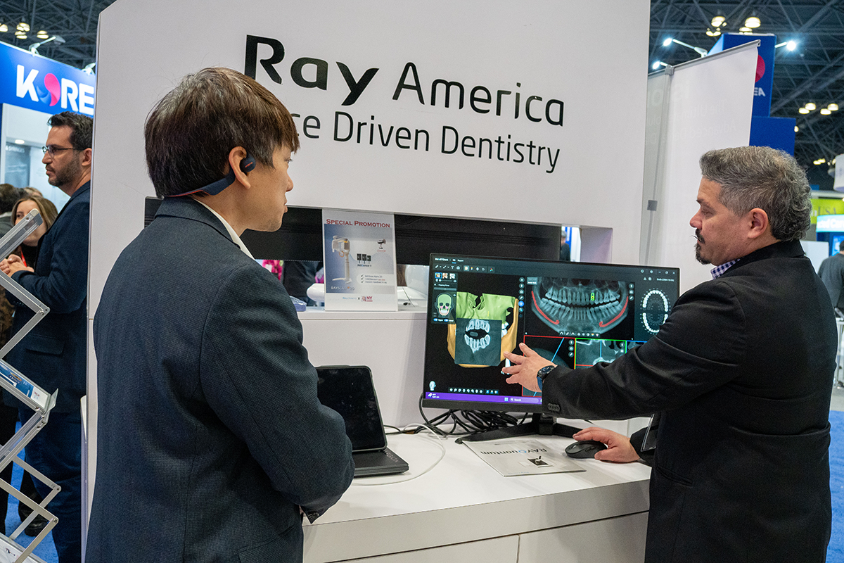 Two men at a booth with a sign that reads "Ray America: AI-Driven Dentistry". One man, wearing a black blazer and a checkered shirt, is pointing at dental images on a computer monitor. The other man, in a dark blazer, listens and observes the screen.