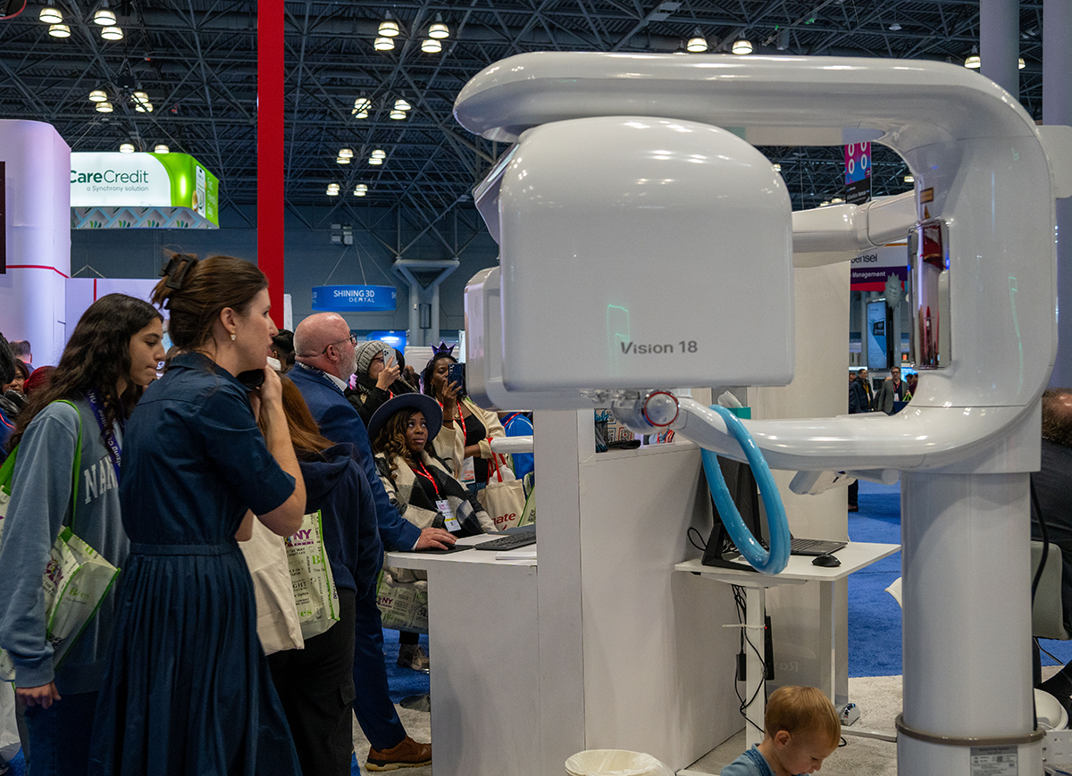 People attending a medical imaging exhibition are gathered around a large white panoramic X-ray machine labeled 'Vision 18' in a spacious convention center.