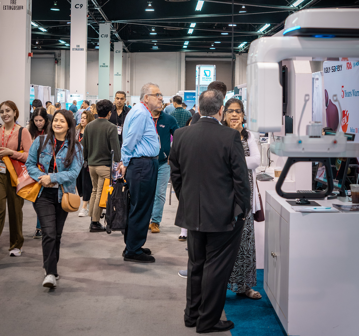 People attending a trade show or exhibition, engaging at booths and displays, with some individuals having conversations and carrying bags, in a large indoor convention center.